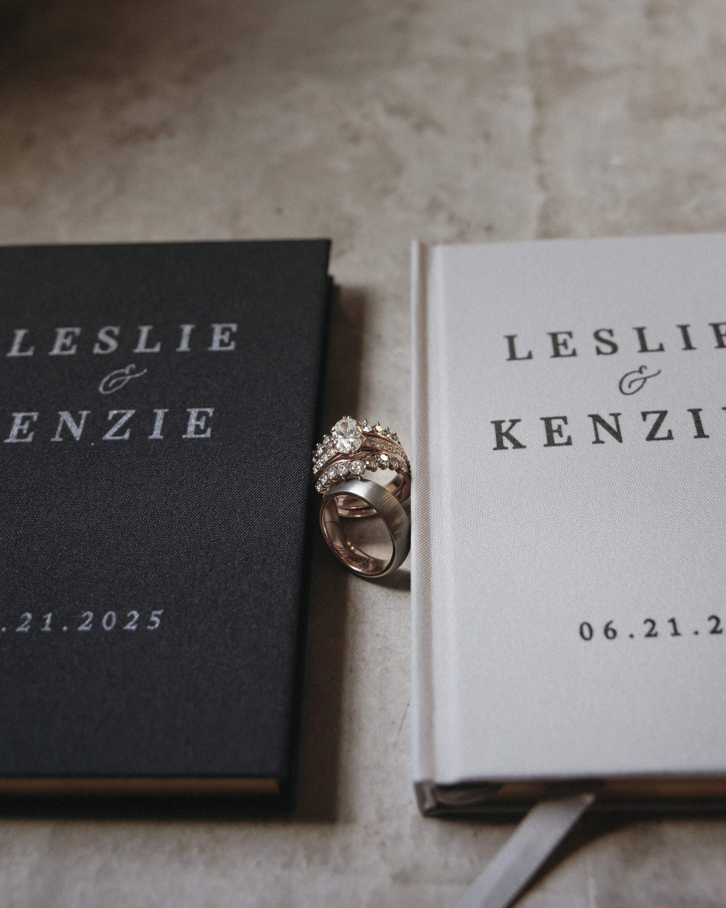 Wedding rings placed between two books titled 'Leslie & Kenzie' with the date 06.21.2025, on a marble surface.