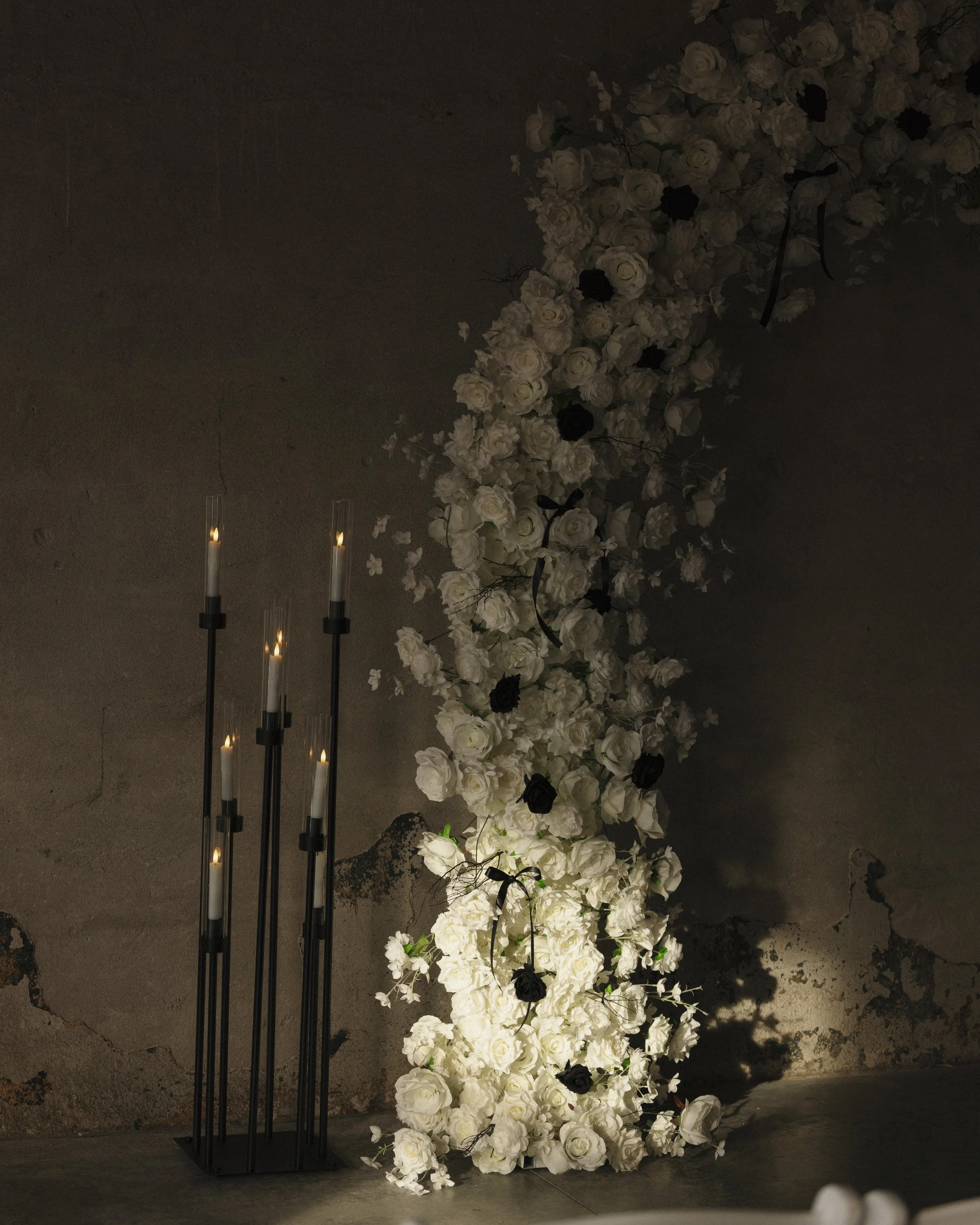 A cascading floral arrangement of white and black roses next to tall black candle holders with lit candles against a textured gray wall.