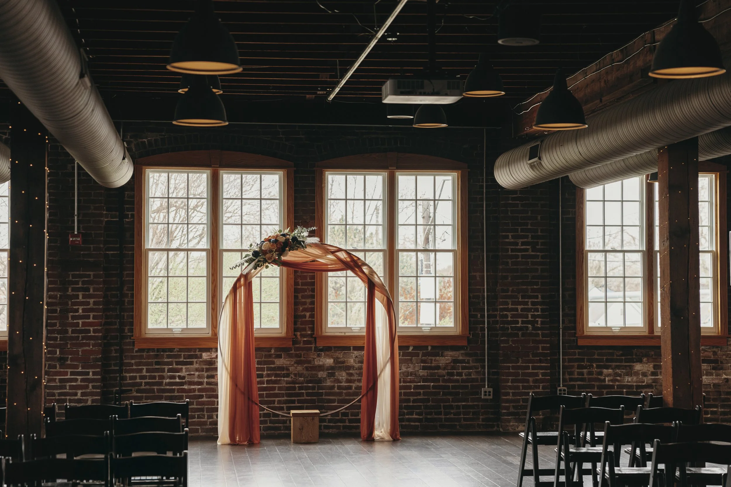 Indoor wedding ceremony setup with a decorated arch in front of large windows, chairs arranged on either side, in a rustic brick-walled venue with hanging pendant lights.