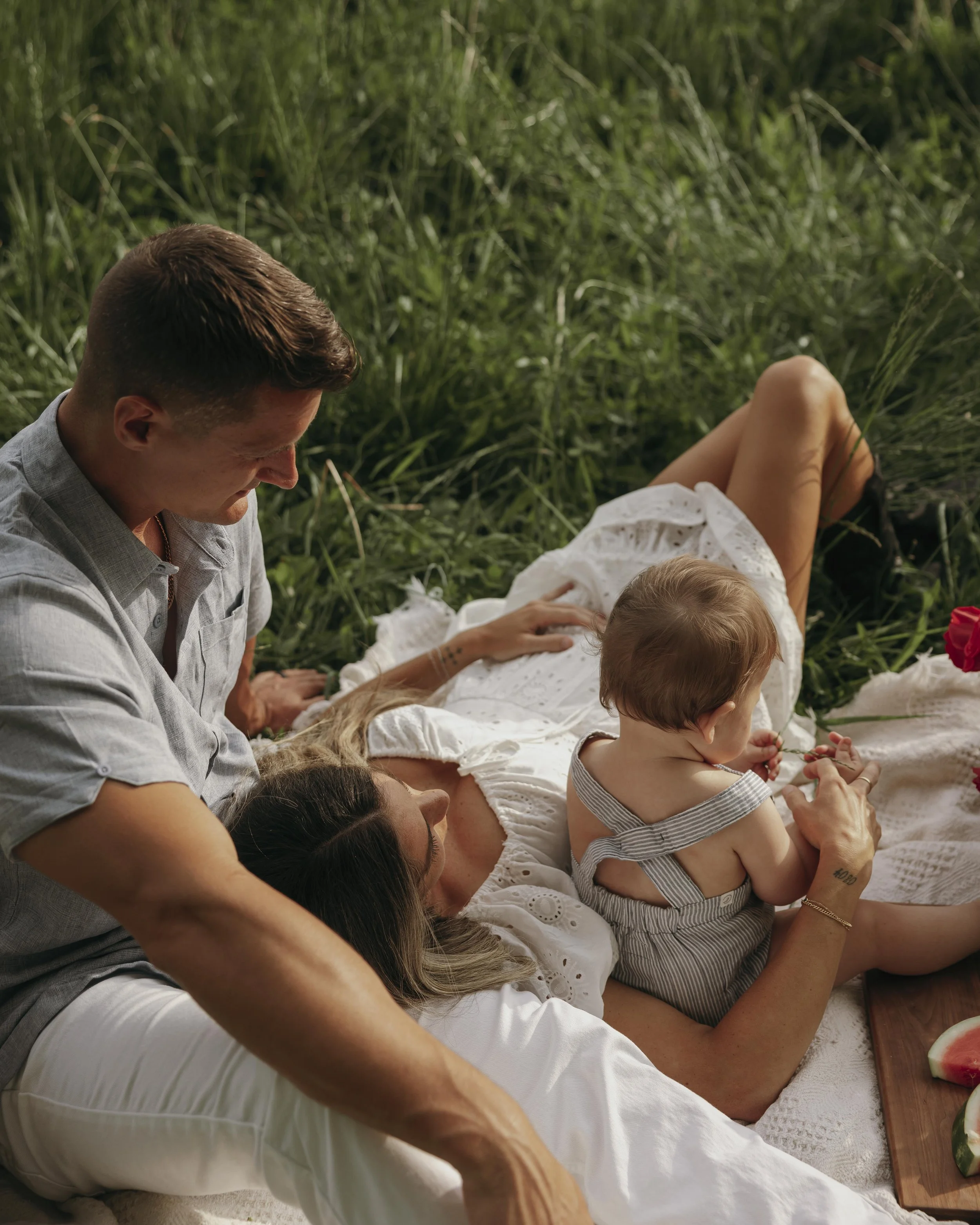 Family having a picnic outdoors on a grassy field, with a woman lying down, a man leaning over her, and a small child sitting on the woman's chest, holding a flower.