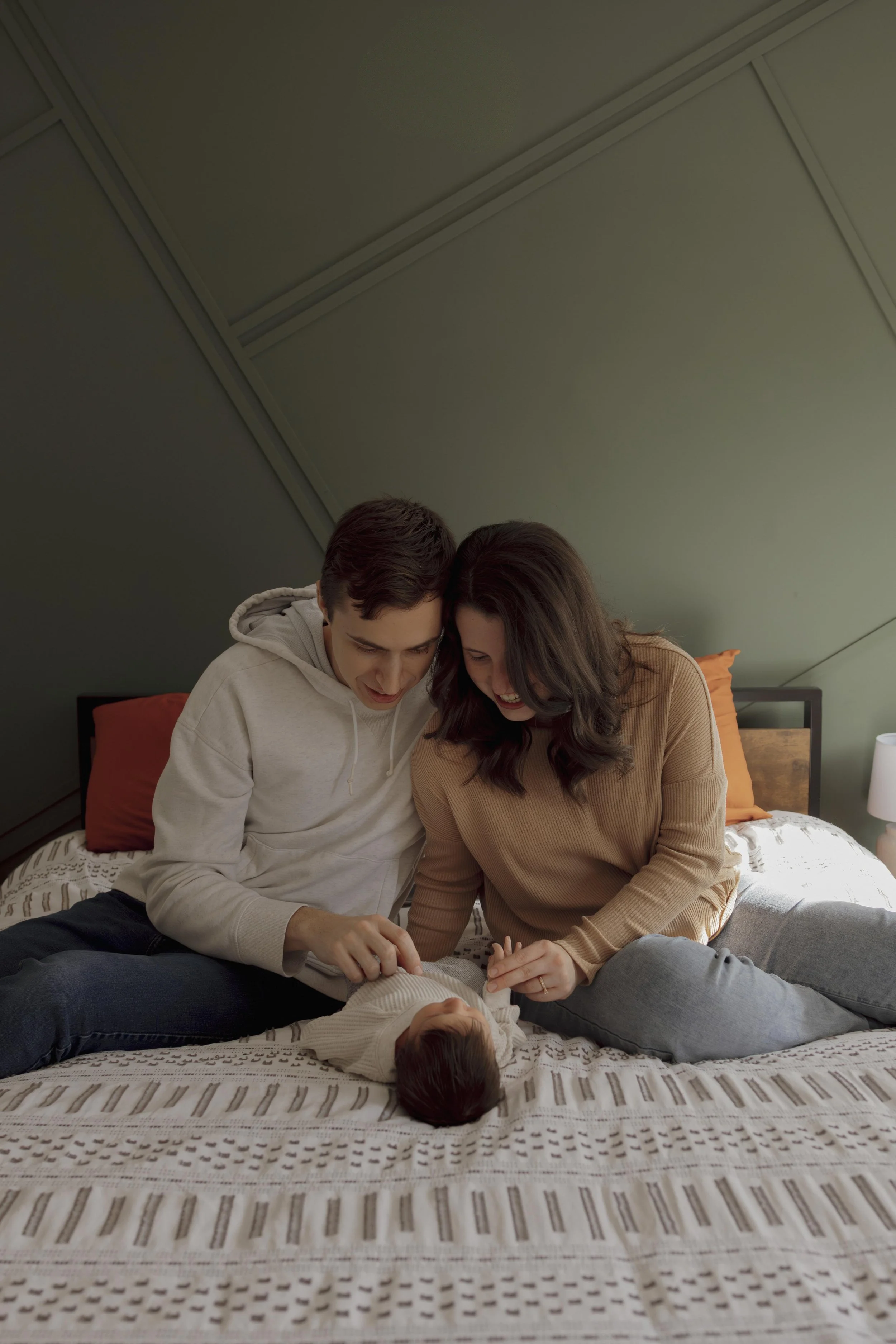 A young couple and their infant lying on a bed, smiling and looking at the baby.