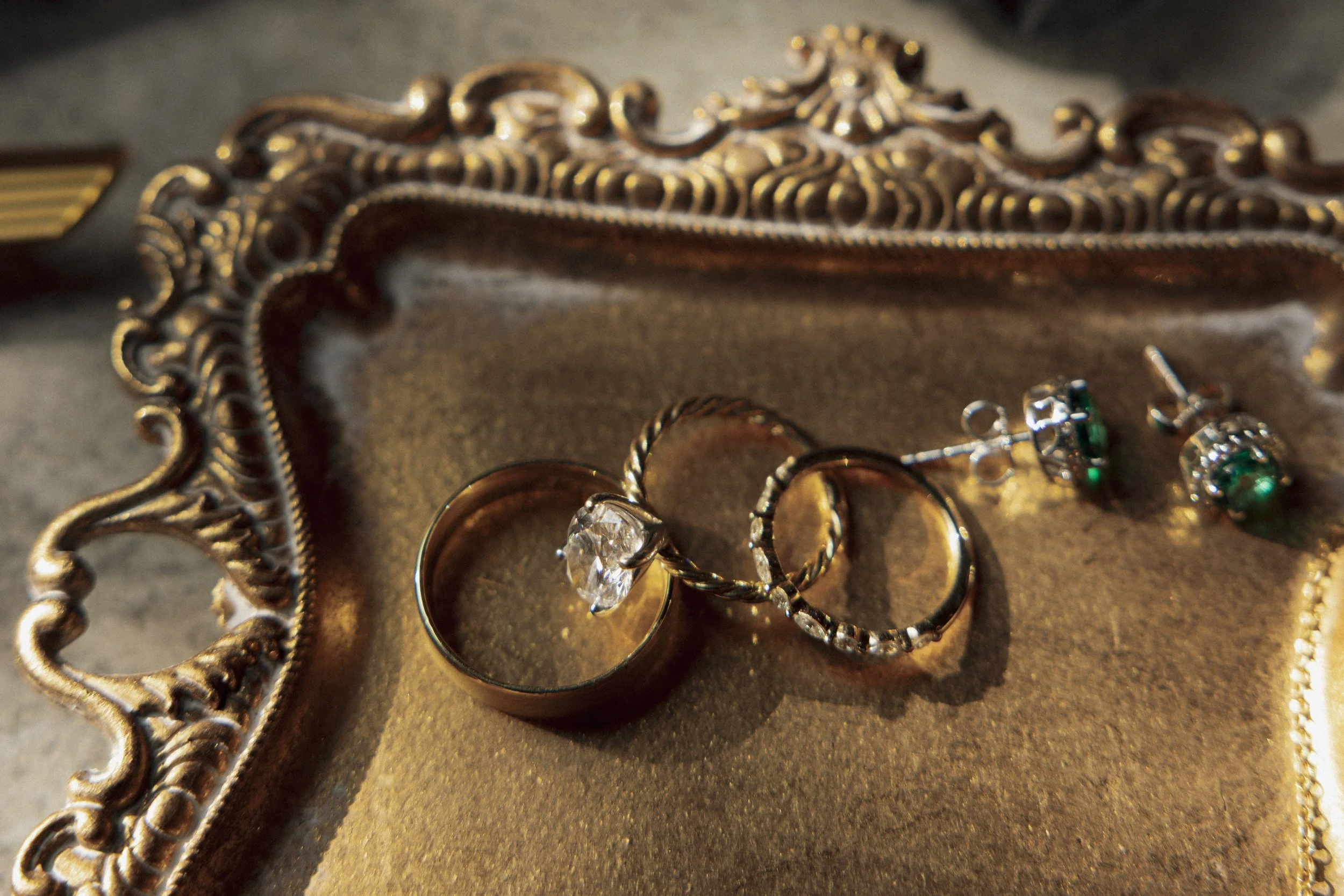 Gold decorative jewelry tray holding a silver ring with a large gemstone, a gold ring, and a pair of earrings with green gemstones.