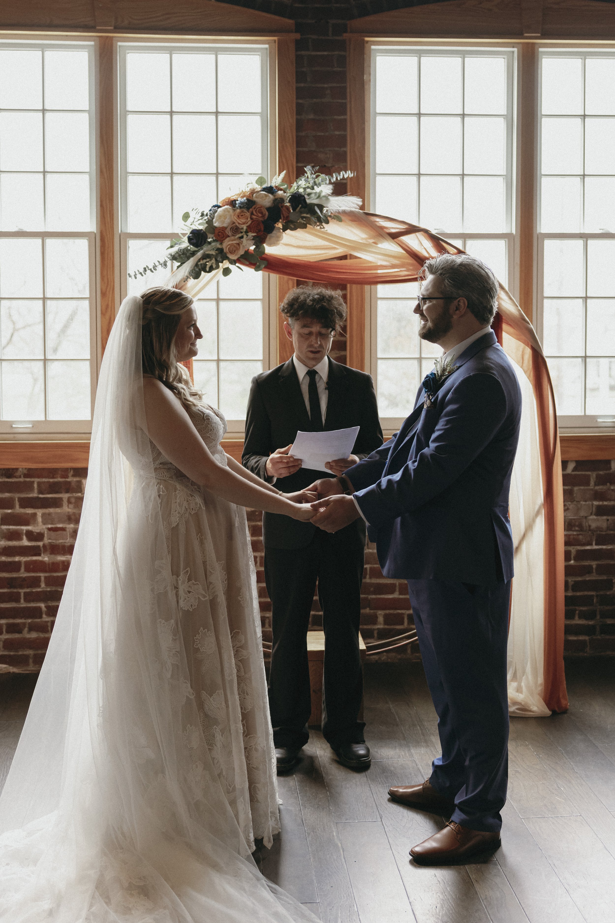 A bride and groom holding hands during their wedding ceremony, with an officiant reading vows, inside a room with brick walls and large window with natural light, decorated with flowers and fabric arch.