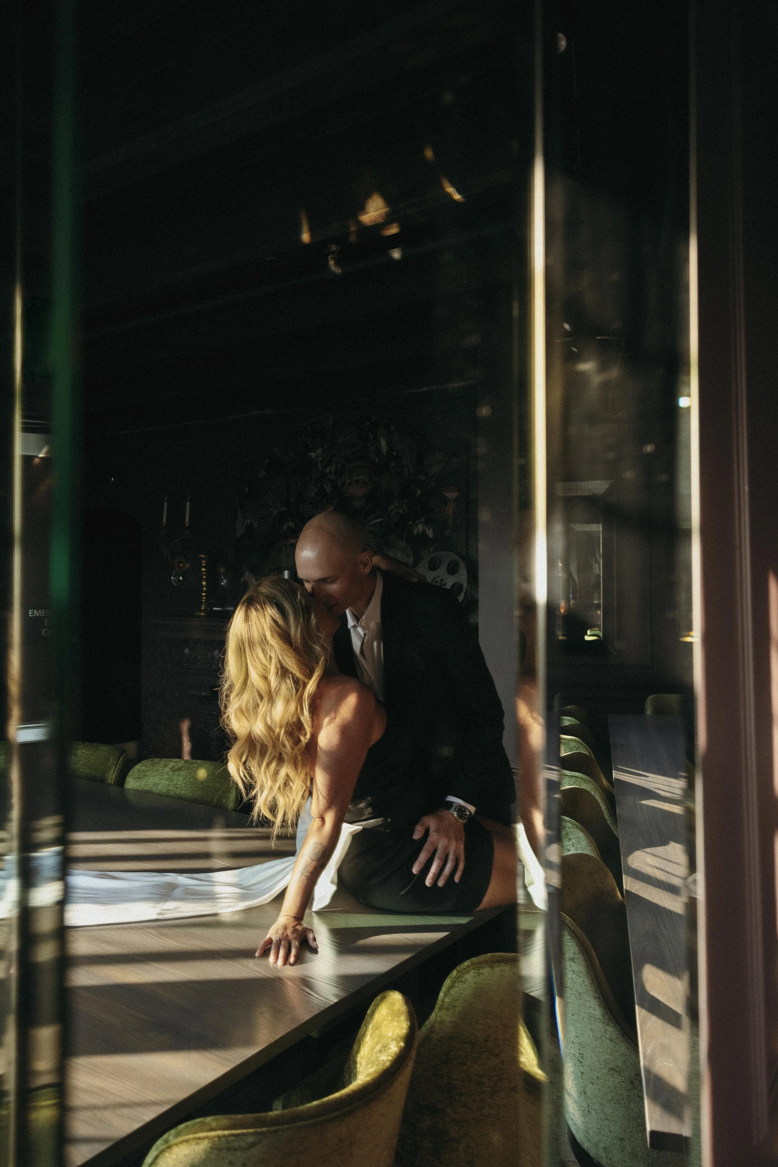 A man and woman share an intimate moment at a bar or restaurant, with the woman leaning on a table and the man leaning close to her, through a glass window.