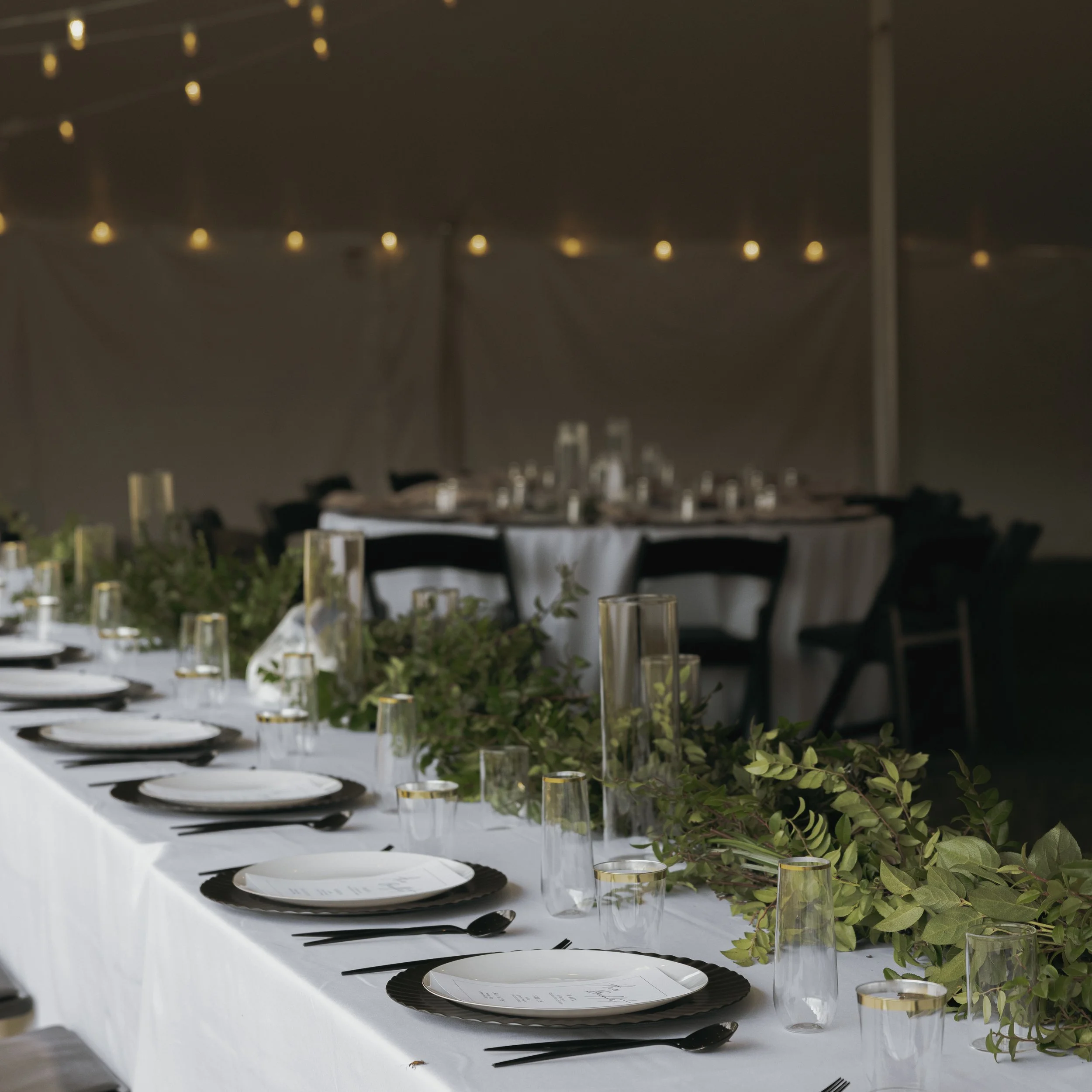 Long banquet table decorated with greenery, vases with candles, plates, and black utensils, set for a formal event under string lights in a tent.