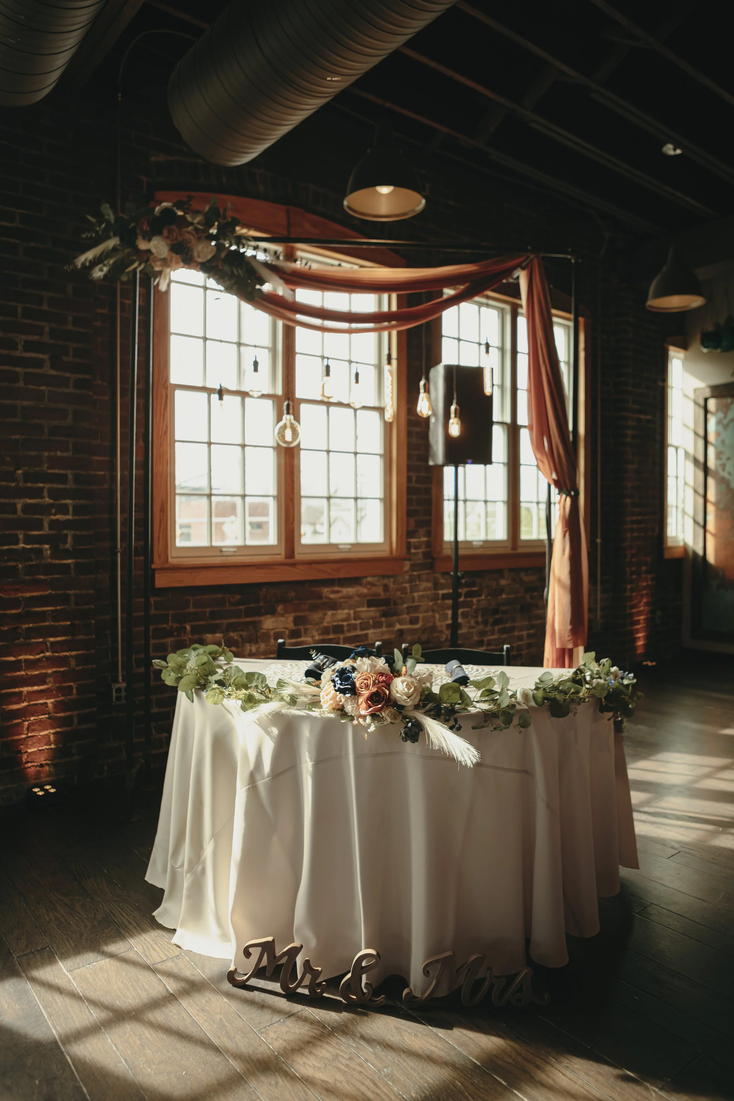 Table with a floral centerpiece and signs reading "Mr & Mrs" in a decorated wedding reception space with brick walls, large windows, and hanging lights.