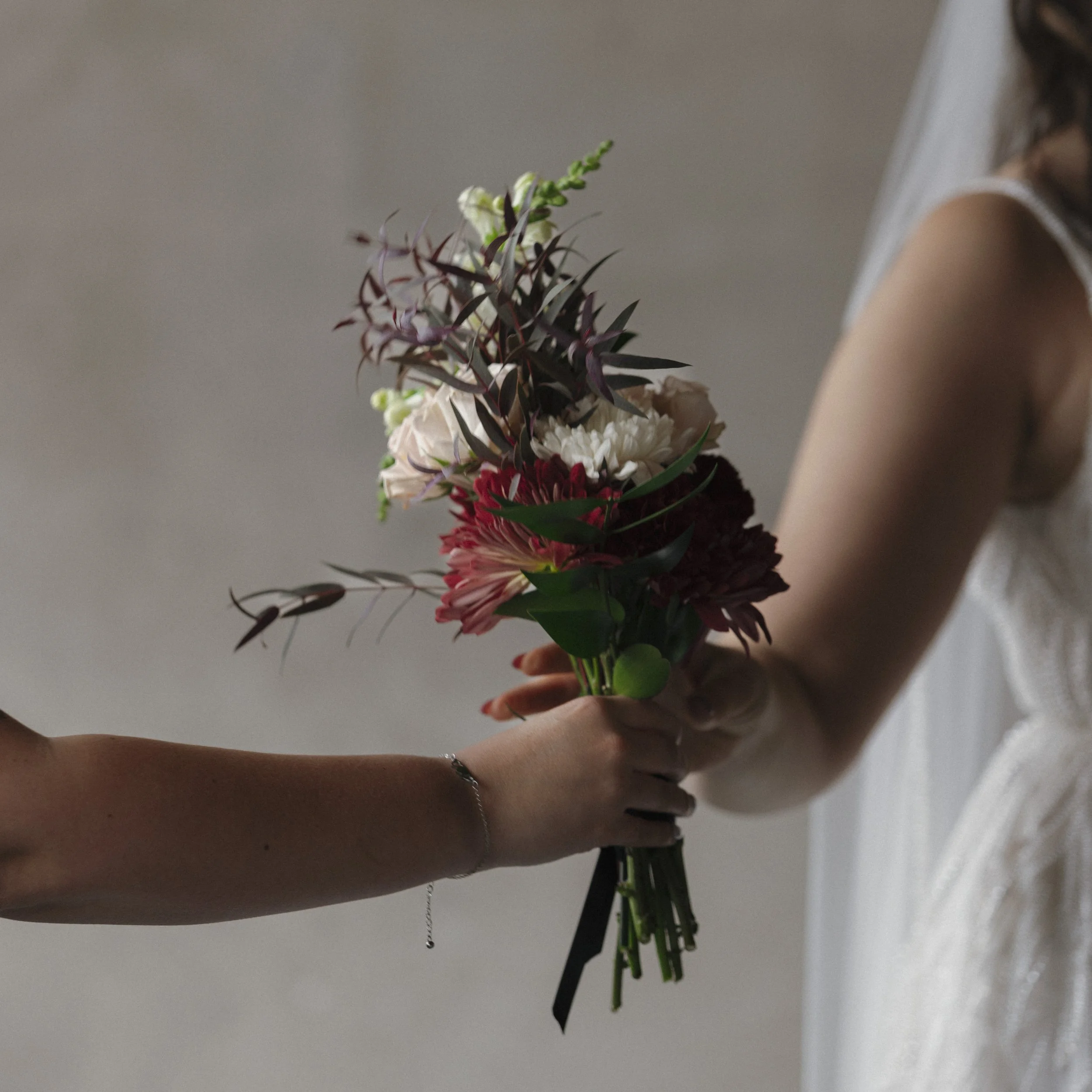 Two women exchanging a bouquet of mixed flowers, including red, white, and purple blooms, against a neutral background.