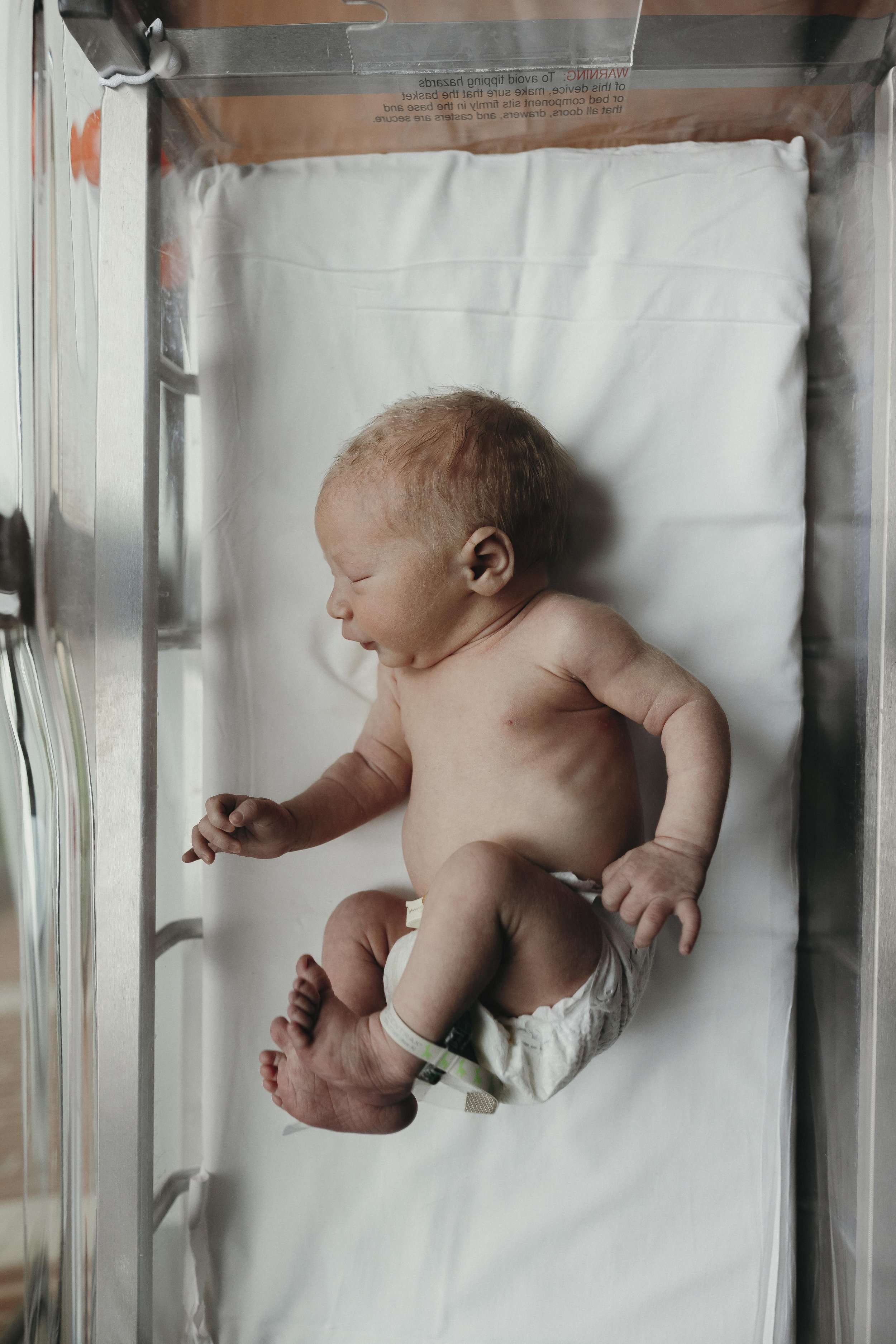 A newborn baby lying on a hospital bassinet mattress with a diaper, seen from above, with a hospital wristband on their ankle.