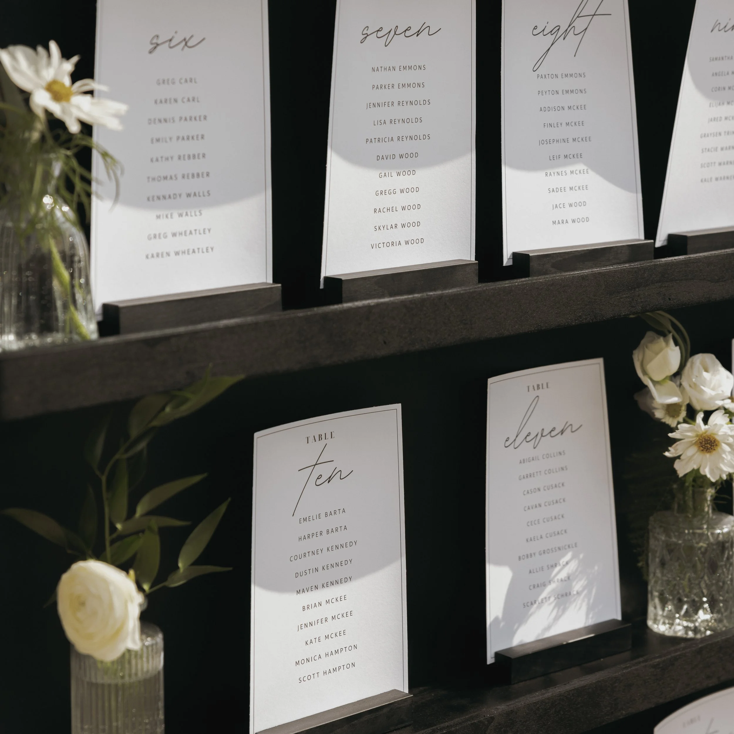 Seating chart with tables numbered six, seven, eight, ten, and eleven, displaying guest names; decorated with white flowers in glass vases.