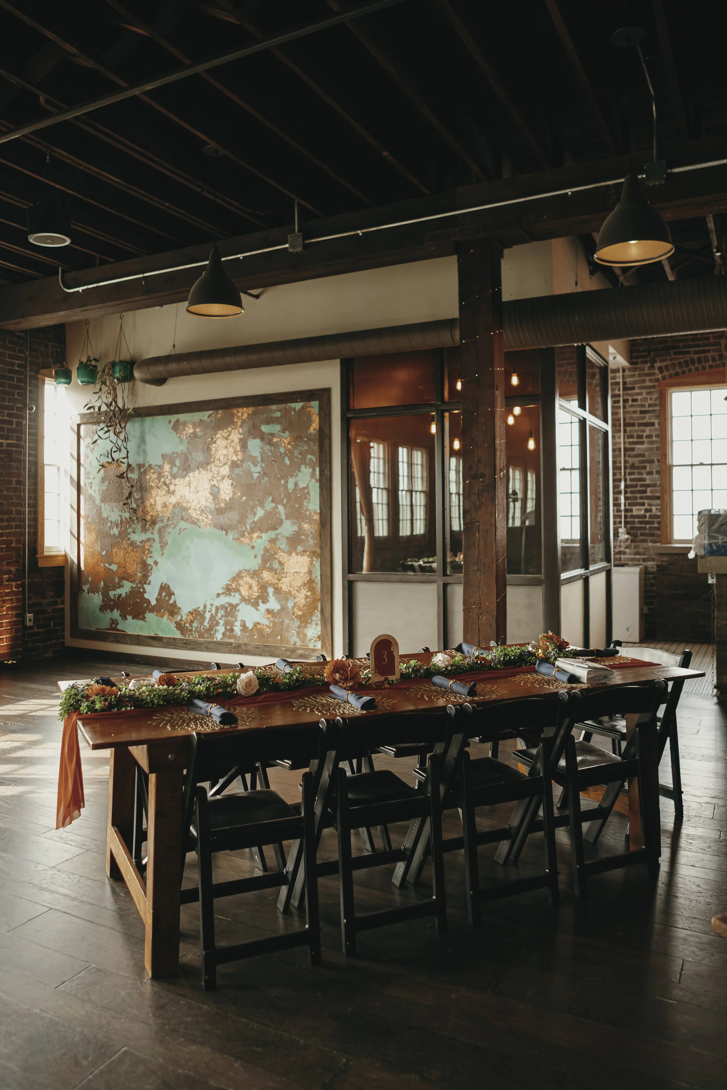 A decorated table set for a wedding reception or special event in an industrial-style venue with brick walls, large windows, and hanging pendant lights.
