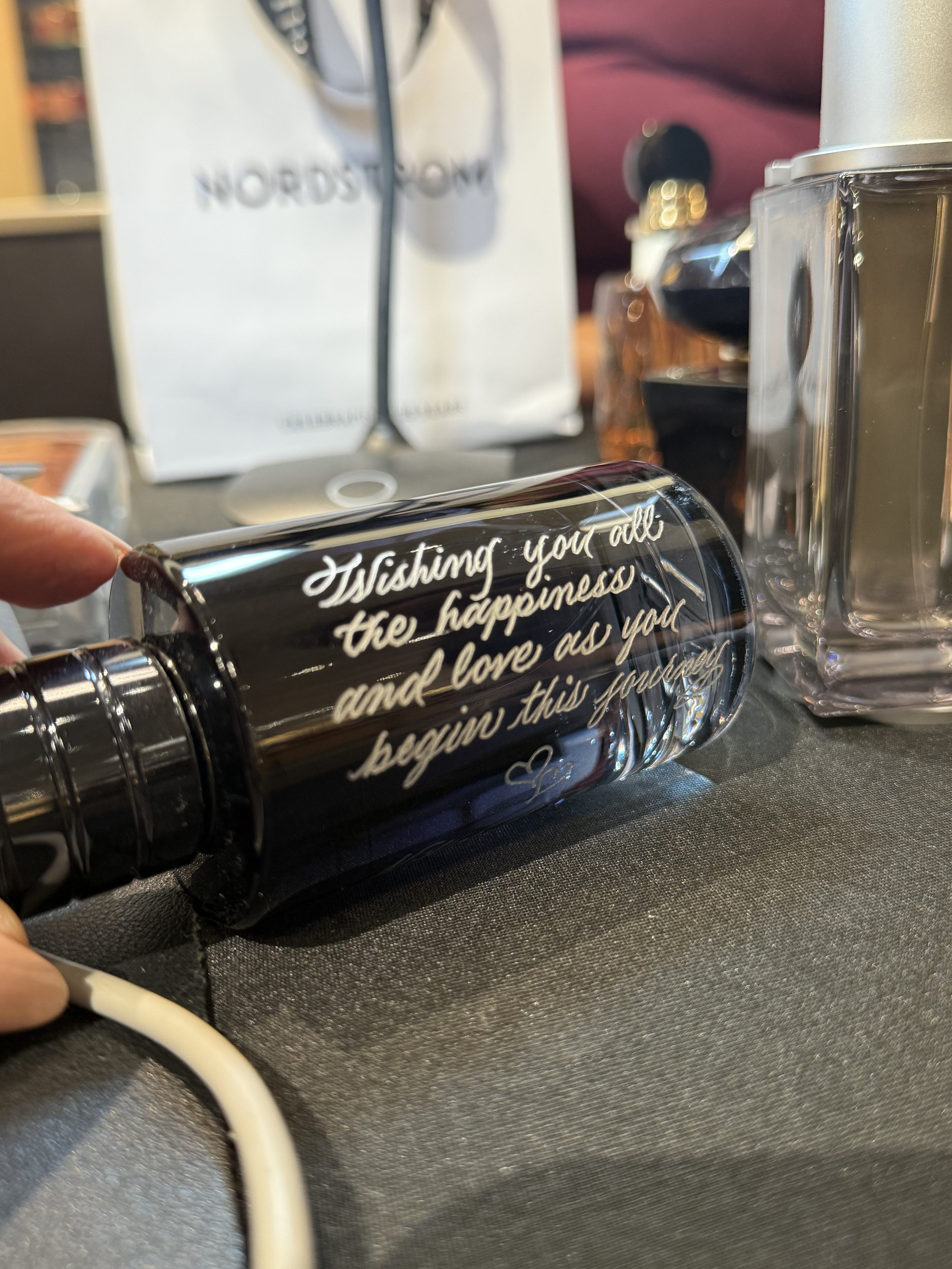 Close-up of a decorative black object with white cursive text that reads, "Wishing you all the true happiness and love as you begin this journey," lying on a black fabric surface. In the background, there is a white Nordstrom shopping bag and several