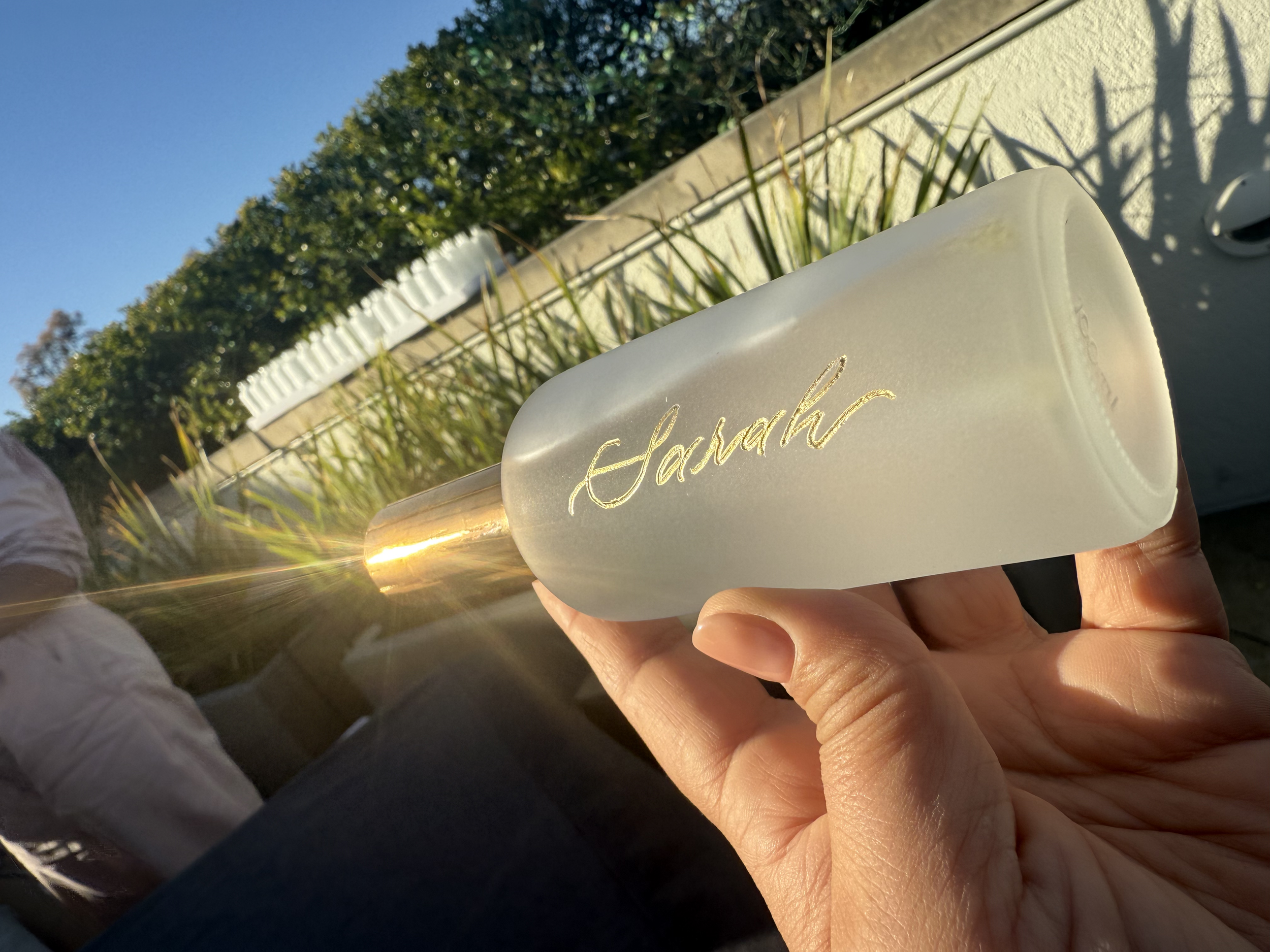 Close-up of a hand holding a white soap bar with the word 'Sarah' written in gold, against a background of blue sky, green bushes, and sunlight.