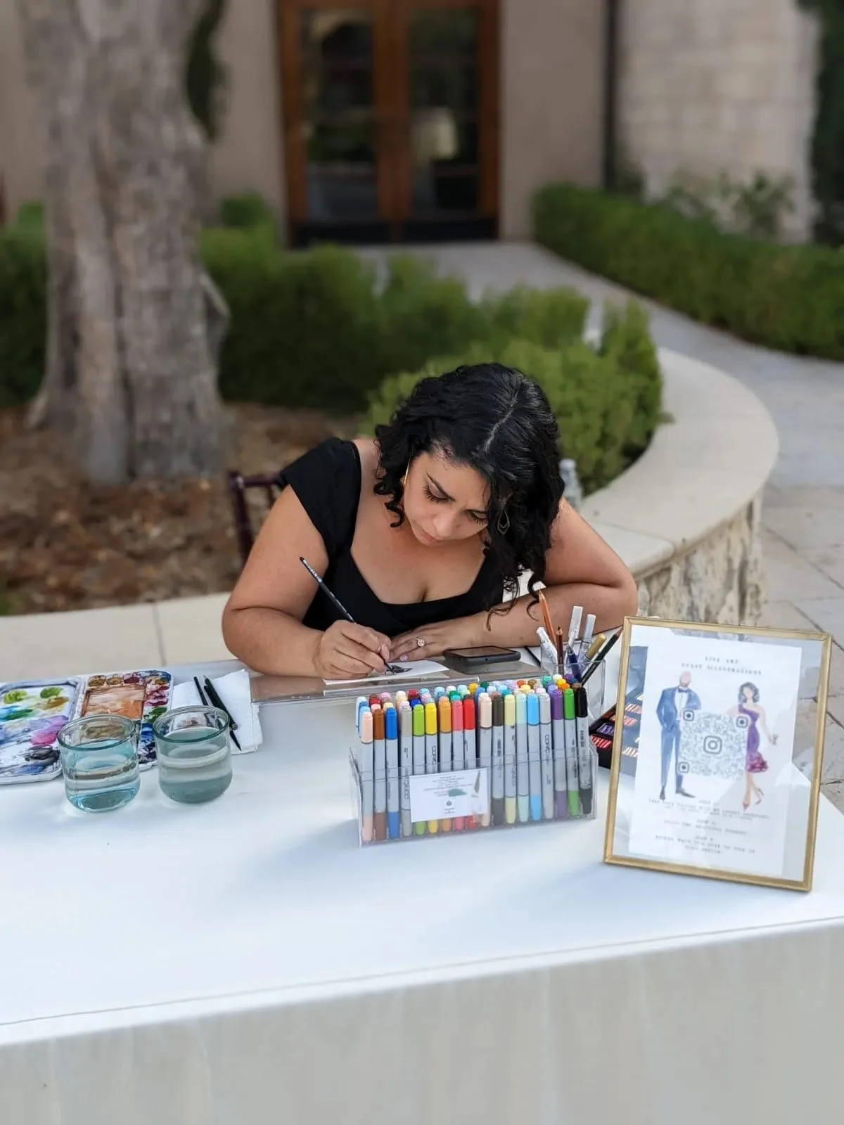 A woman sitting at a table outdoors, coloring with markers, with various art supplies, water glasses, and a framed sign on the table.