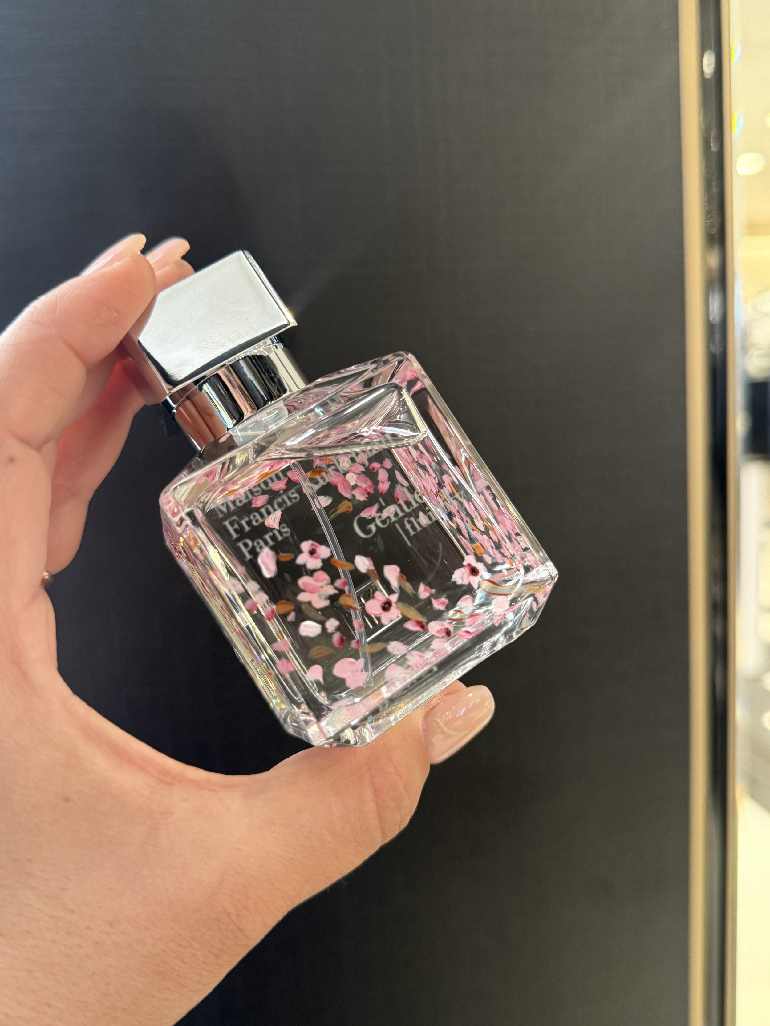 A hand holding a rectangular perfume bottle with a reflective silver cap and a floral design on the glass, featuring pink flowers and black background.