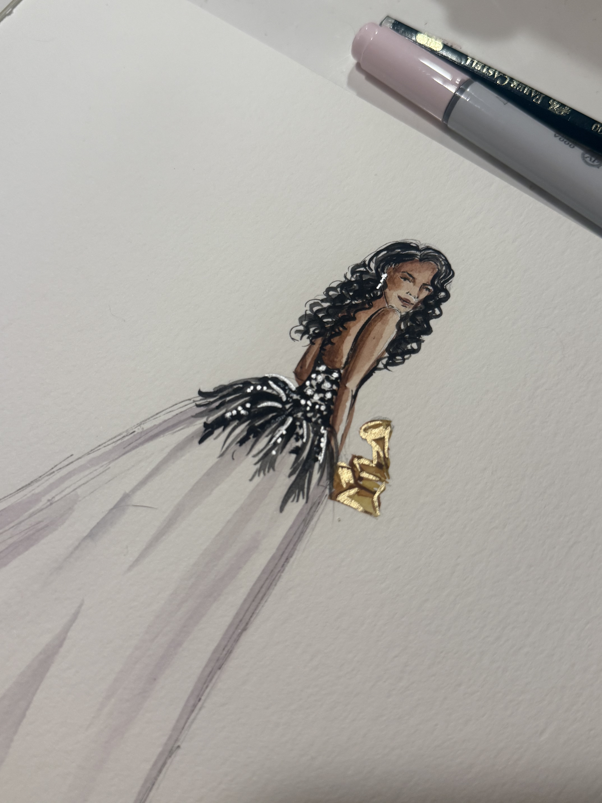 A fashion illustration of a woman with dark, curly hair, wearing a black dress with silver accents, gold boots, and earrings, drawn on white paper with some pens and a pencil nearby.