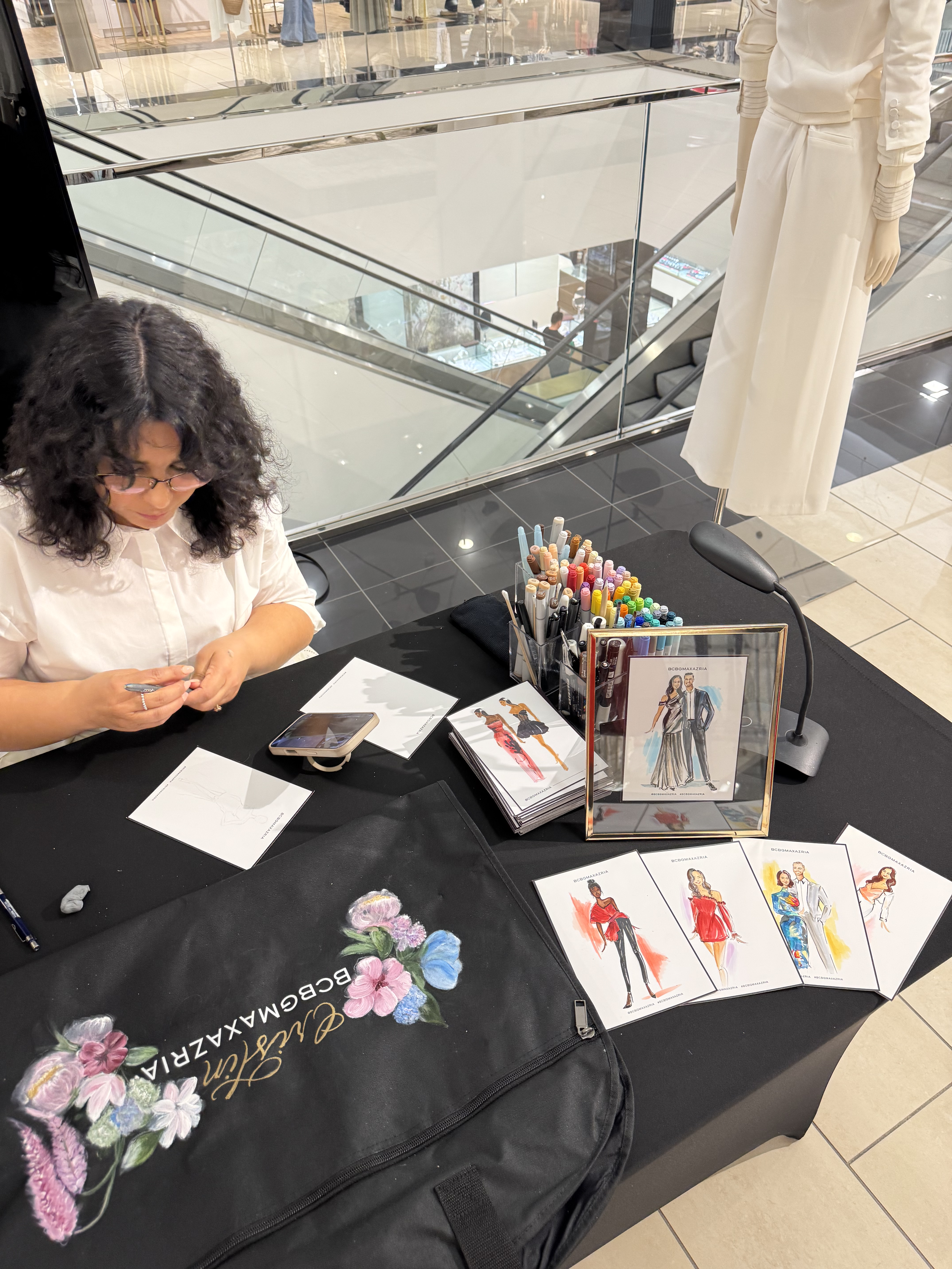 A woman with curly black hair and glasses sitting at a table in a shopping mall, working on sketches of fashion designs featuring models in various outfits, surrounded by colored markers and fashion illustrations.