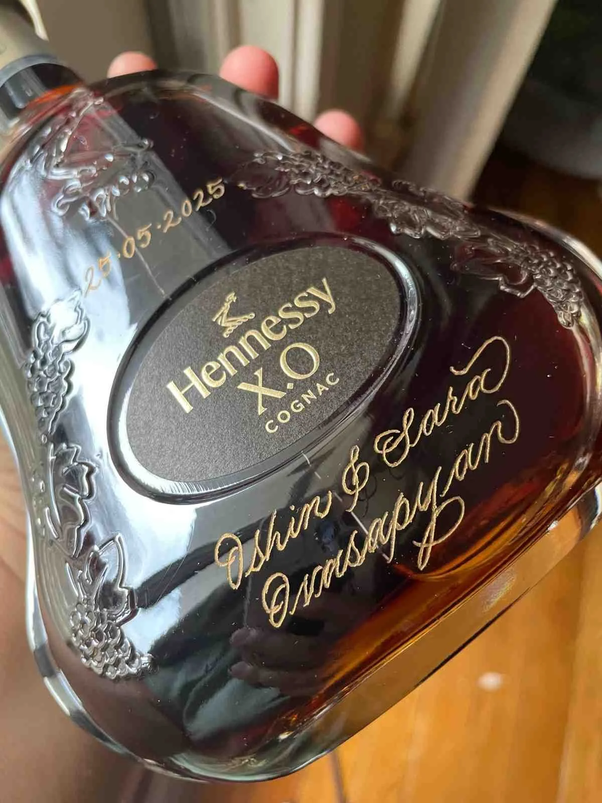 A clear glass bottle of Hennessy XO Cognac with ornate gold lettering and decorative patterns, resting on a surface.