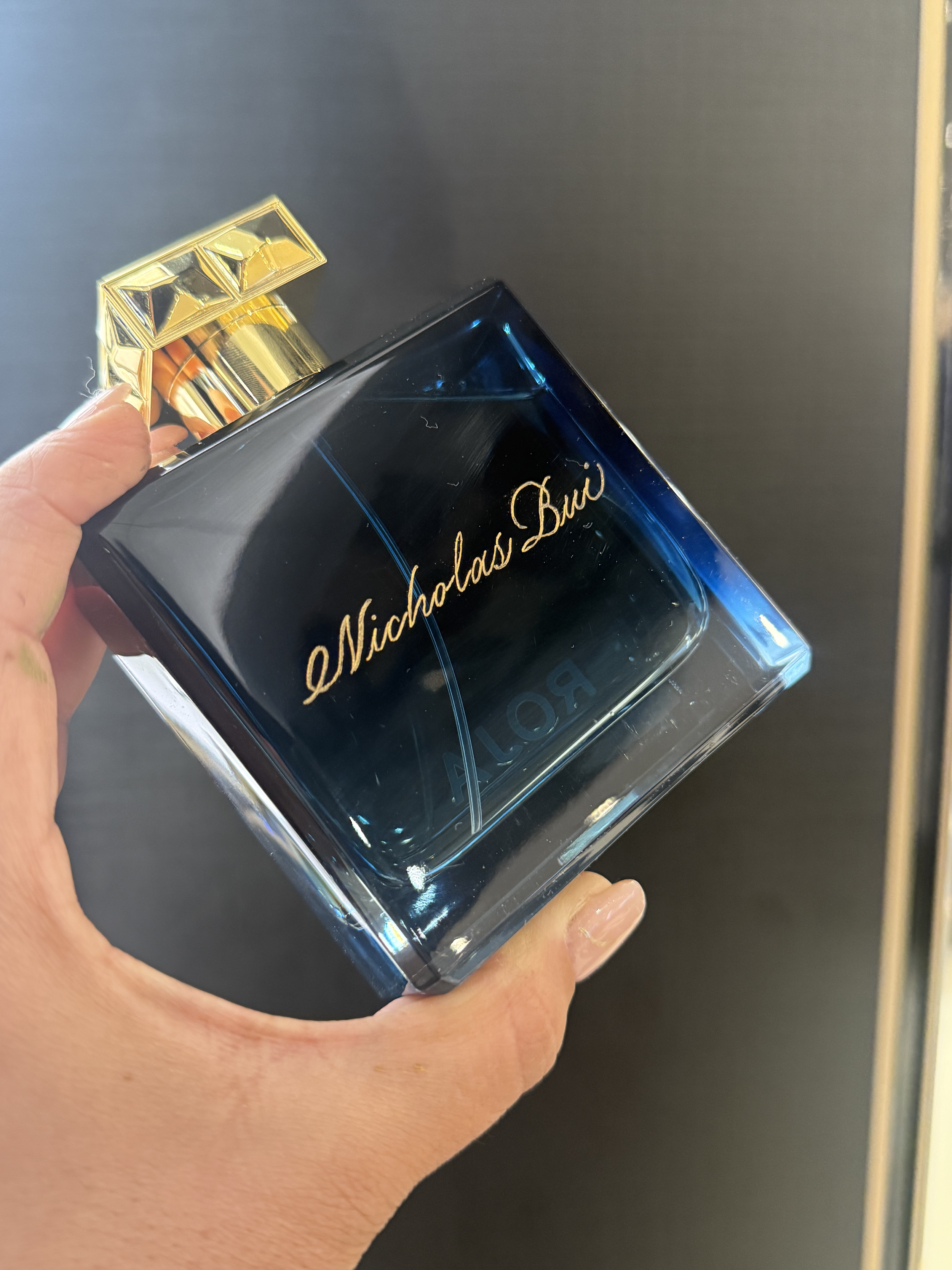 Close-up of a hand holding a perfume bottle labeled 'Nicholas Buzz' with a gold cap against a dark background.