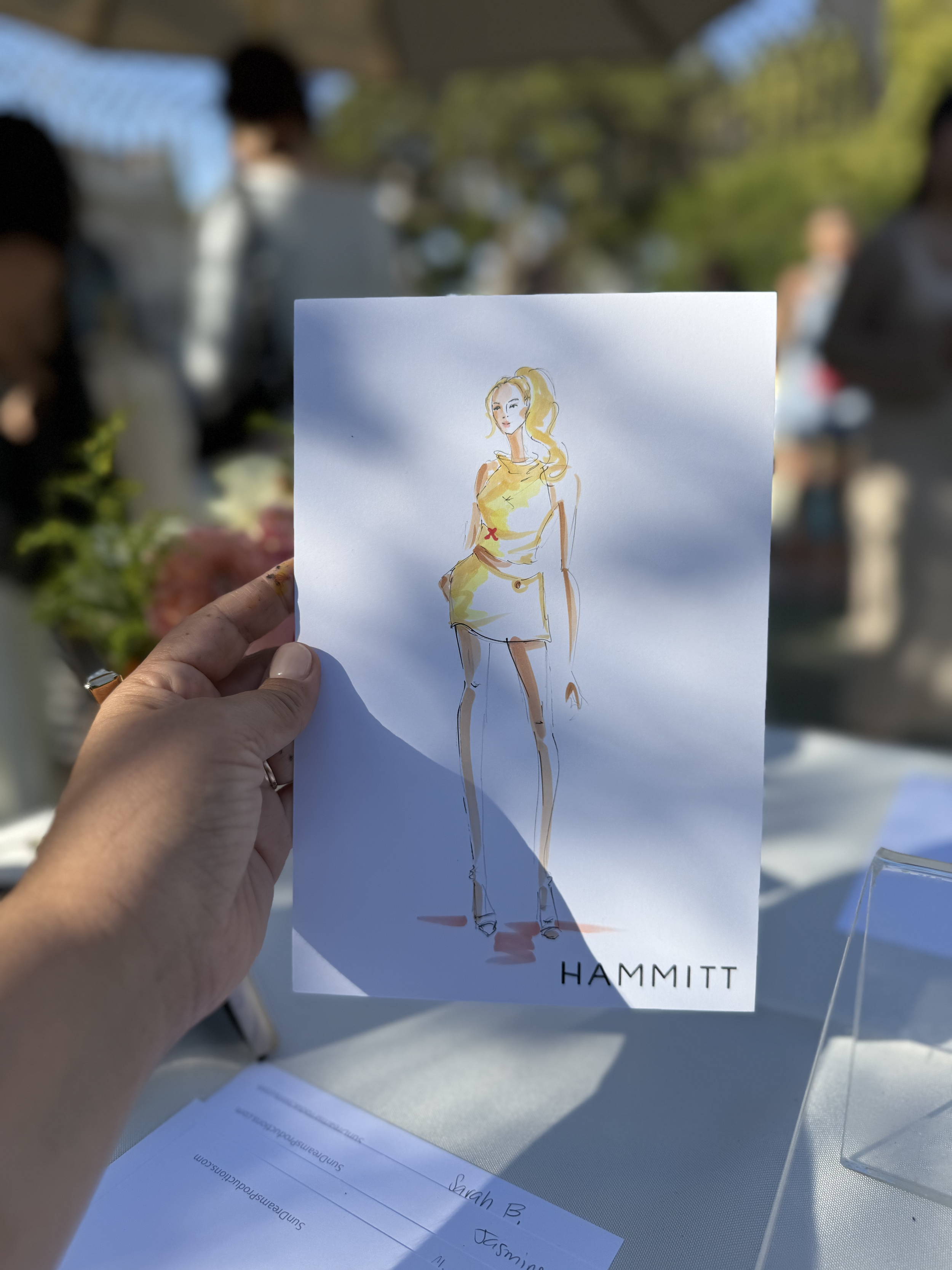 A hand holding a paper with a fashion sketch of a woman in a yellow outfit. In the background, there are blurred figures and trees, indicating an outdoor event.