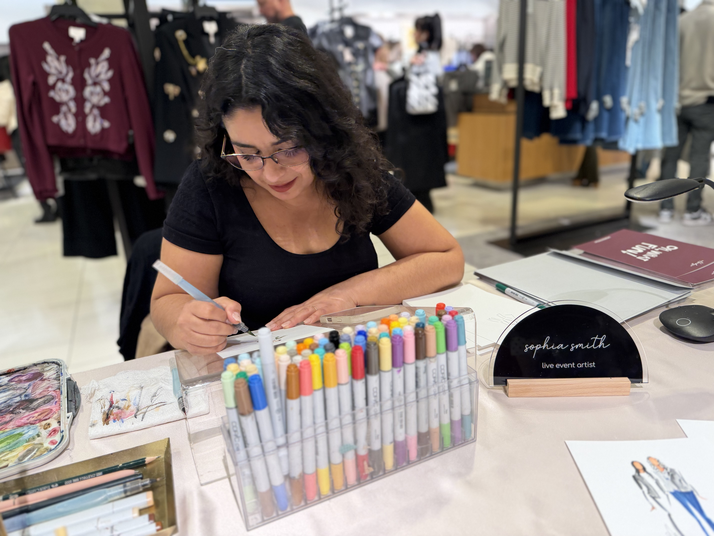 Woman with black curly hair and glasses sitting at a table with art supplies, including paints, markers, and paper, in a retail store with clothing racks in the background.