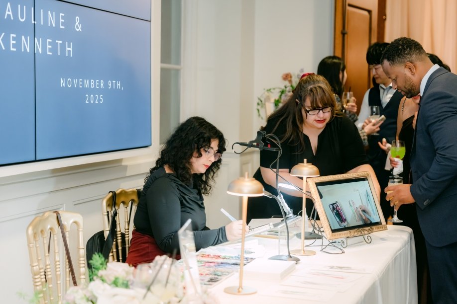Wedding reception registration table with a woman signing in, a tablet displaying a photo, and guests socializing with drinks in hand.