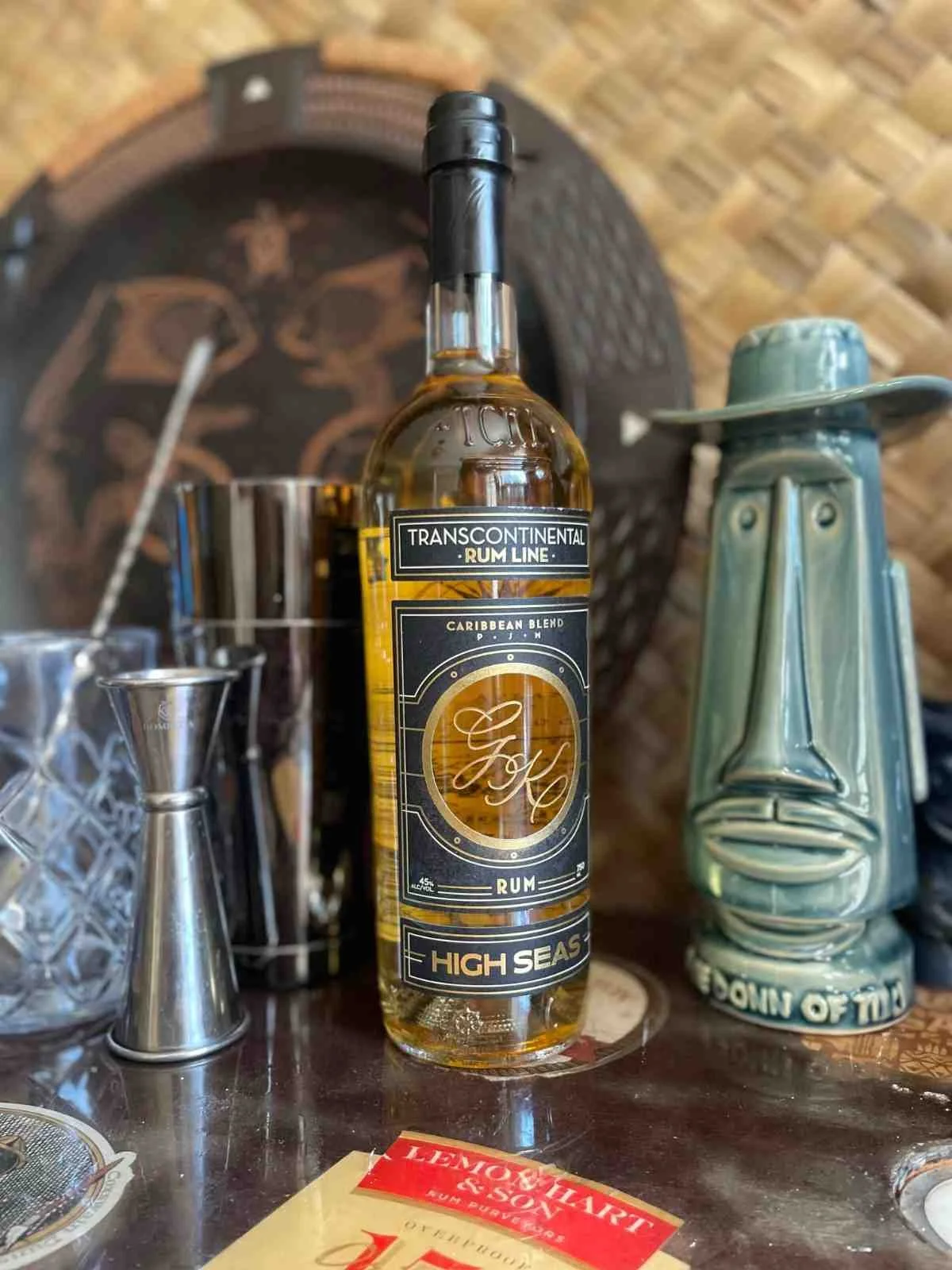 A bottle of Transcontinental Rum Line Caribbean Blend High Seas Rum on a bar counter surrounded by barware and decorative items, including a ceramic face sculpture.