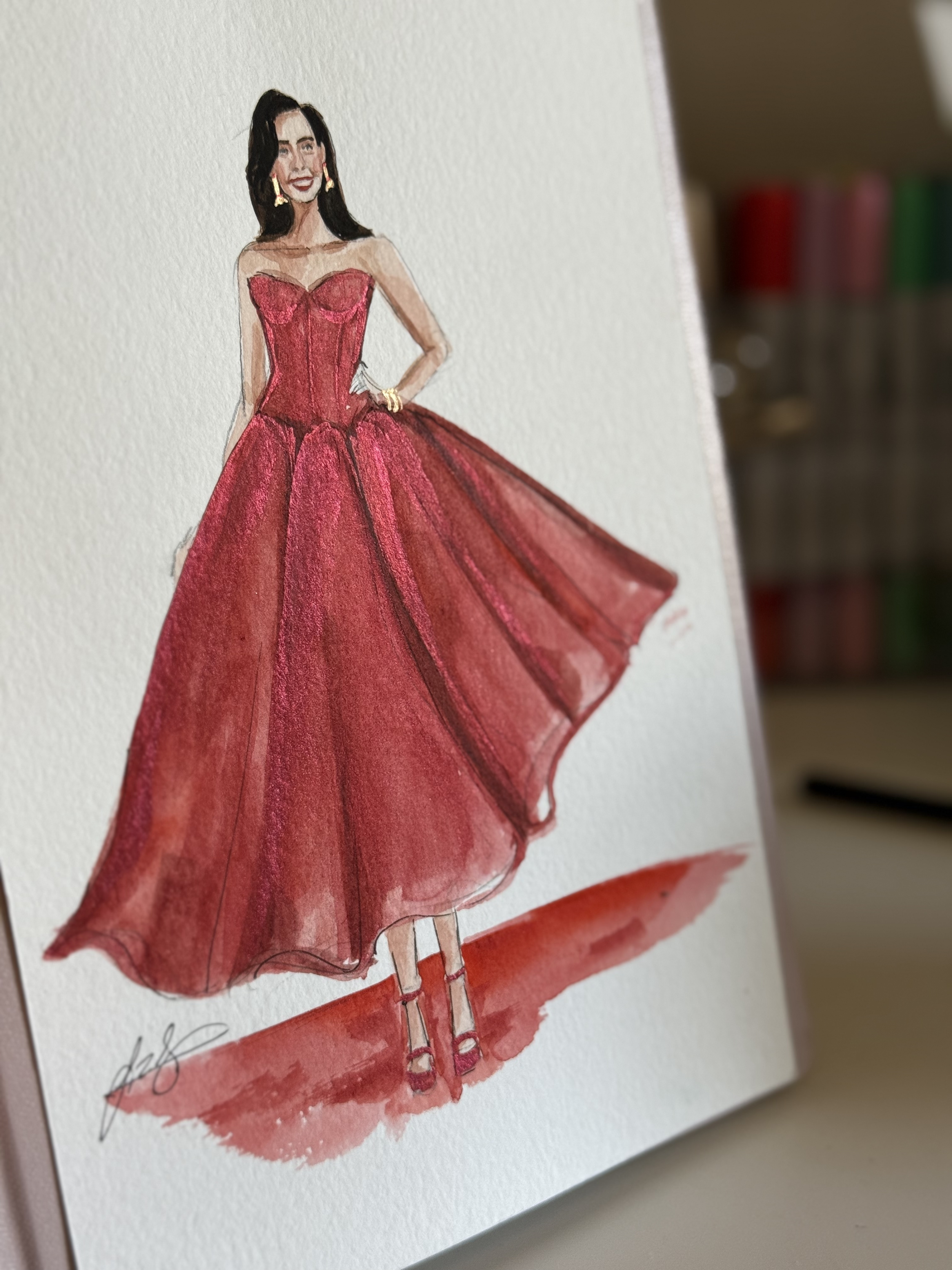 Watercolor fashion illustration of a woman in a strapless red gown with a full skirt, black hair, gold earrings, and a gold bracelet.