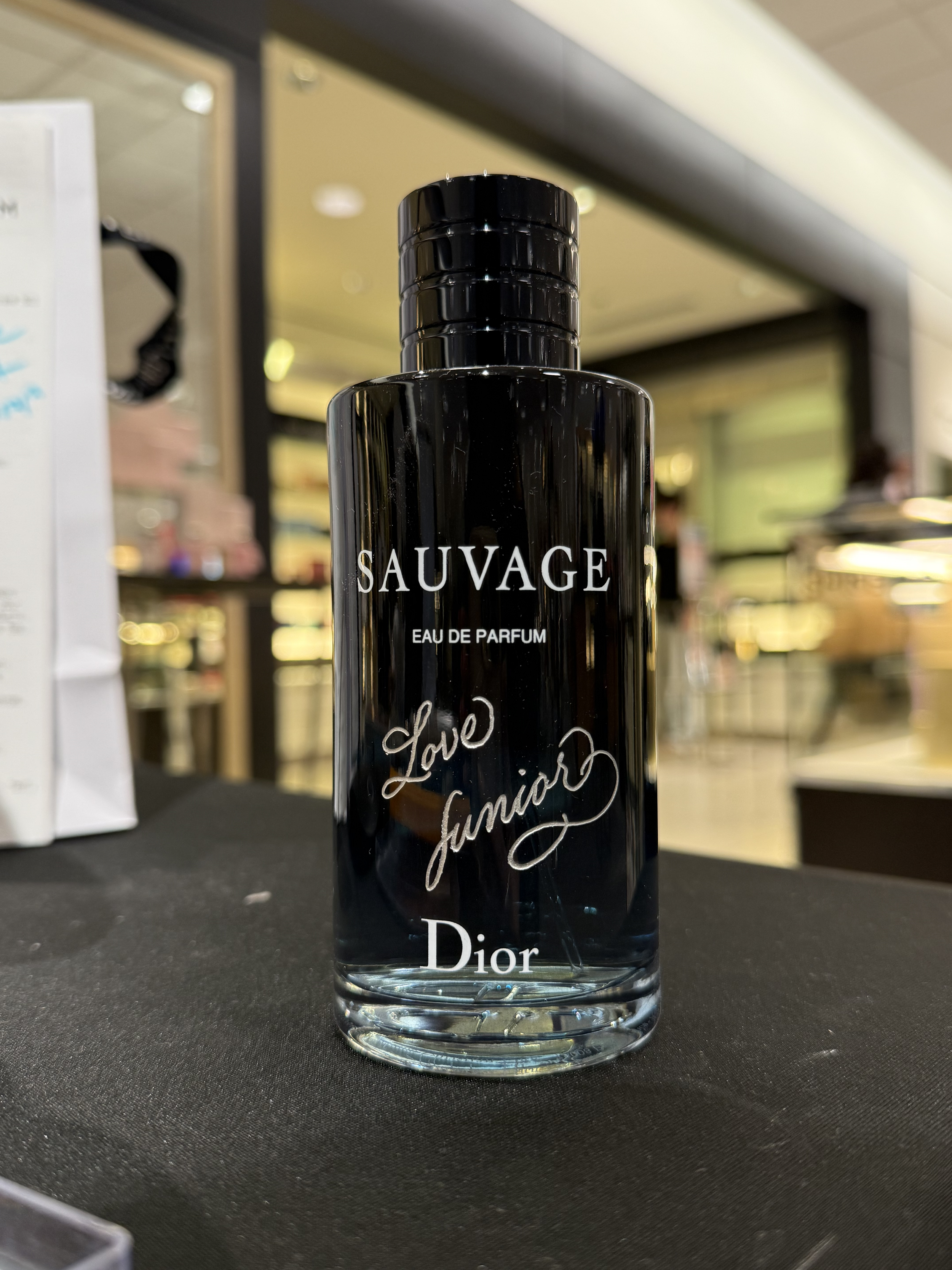 A bottle of Dior Sauvage Eau de Parfum with black glossy finish and cursive writing on a black surface, blurred background of a store or shopping mall.