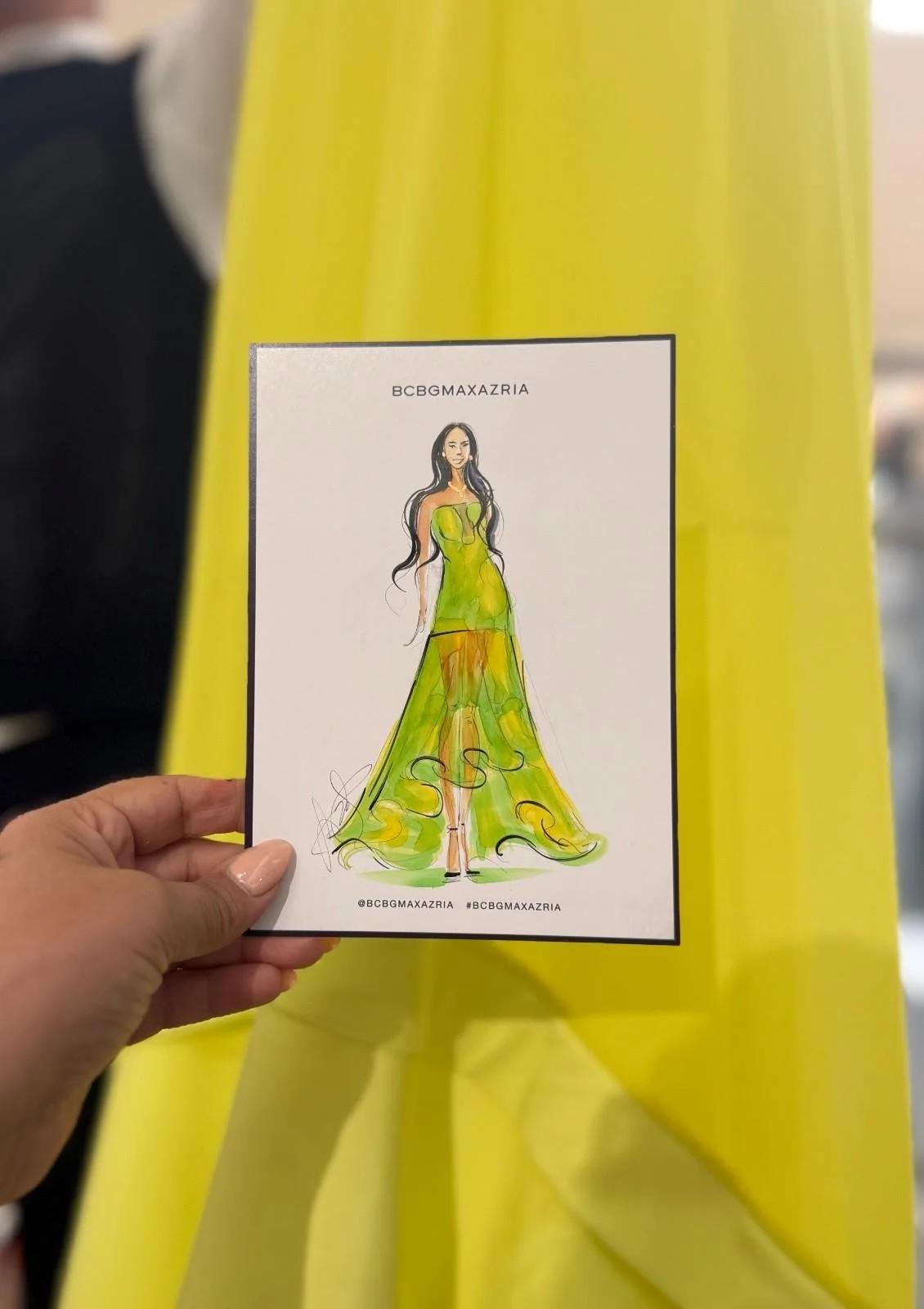 A hand holding a fashion sketch card of a woman in a long, flowing yellow-green dress with black accents, set against a yellow background.