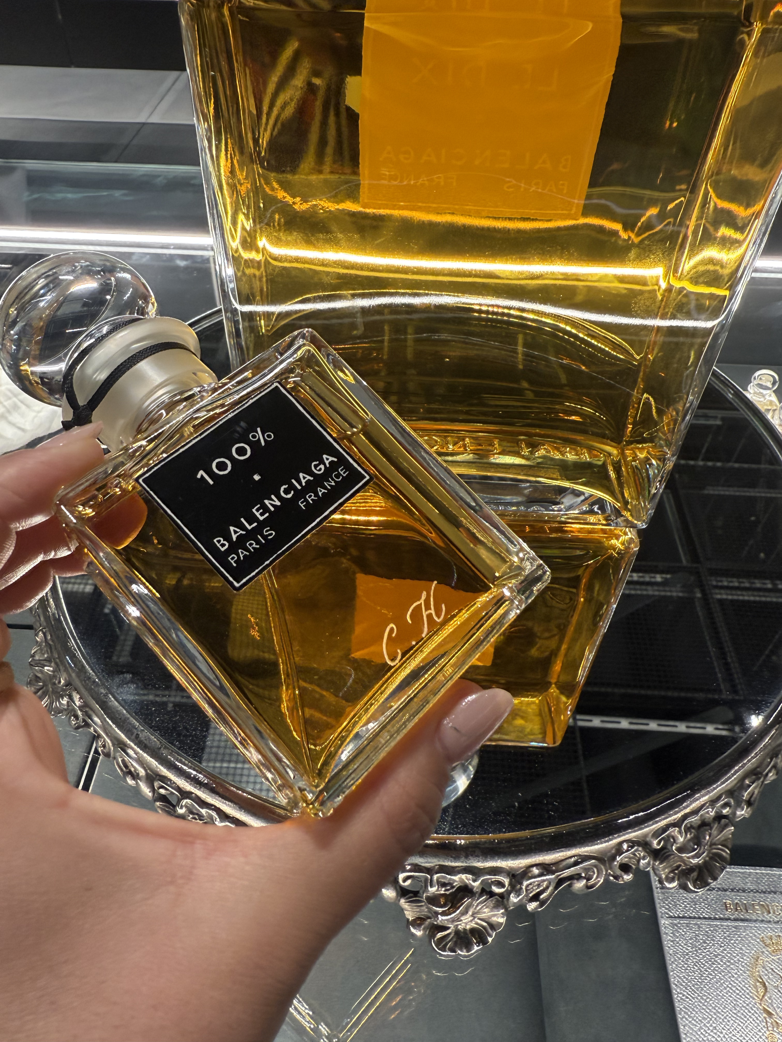A person holding a bottle of Balenciaga Paris perfume in front of a large decorative glass container filled with yellow liquid. The background shows a silver tray, a silver box, and a decorative silver tray with an intricate border.