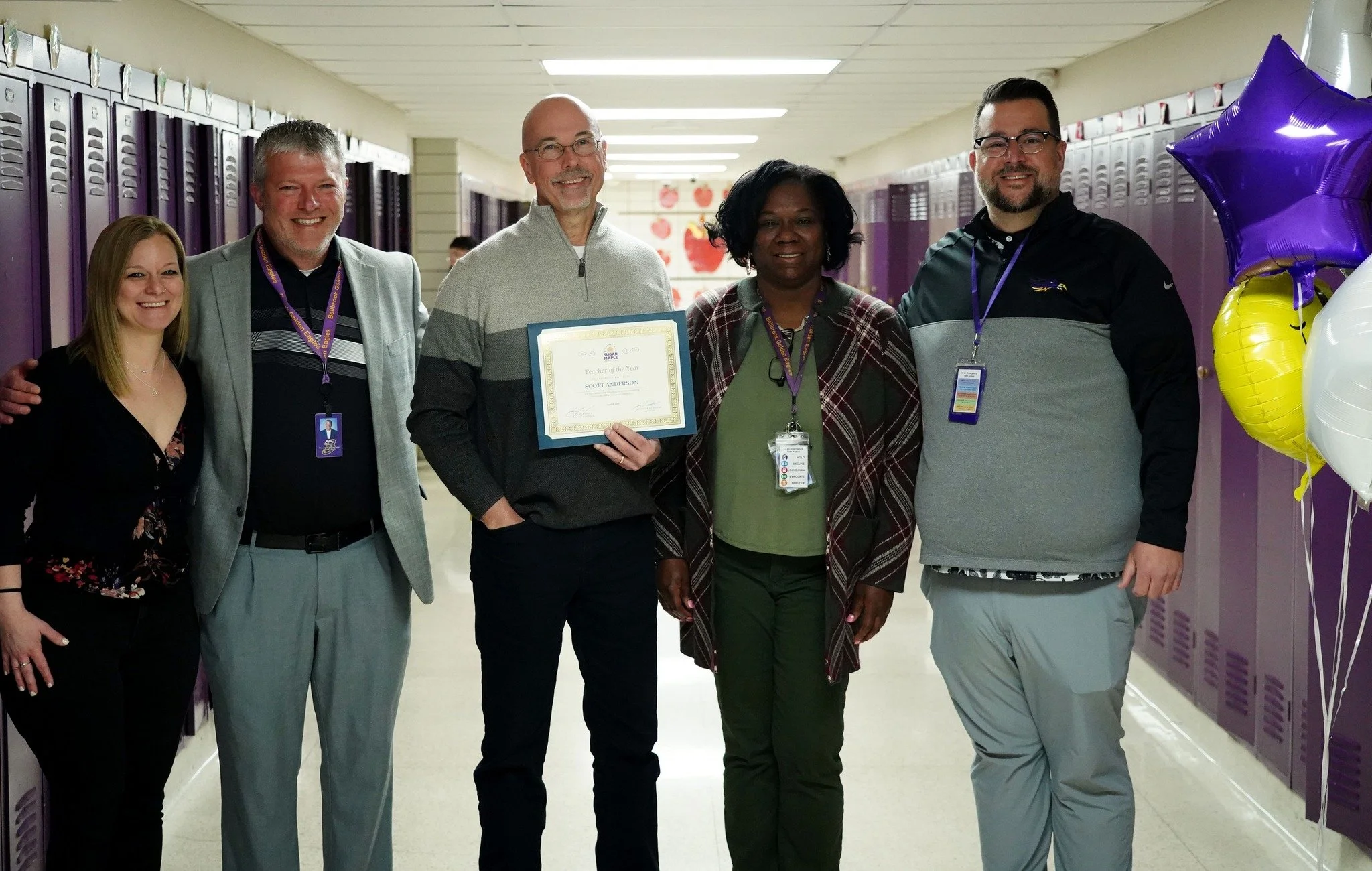 Five people standing together in a school hallway with purple lockers, celebrating an achievement. One person holds a framed certificate.