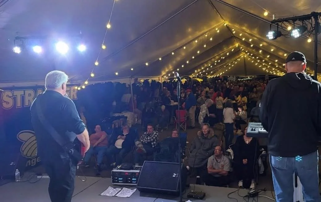 A man speaking on stage at a crowded event under a large tent decorated with string lights, with an audience seated and watching.