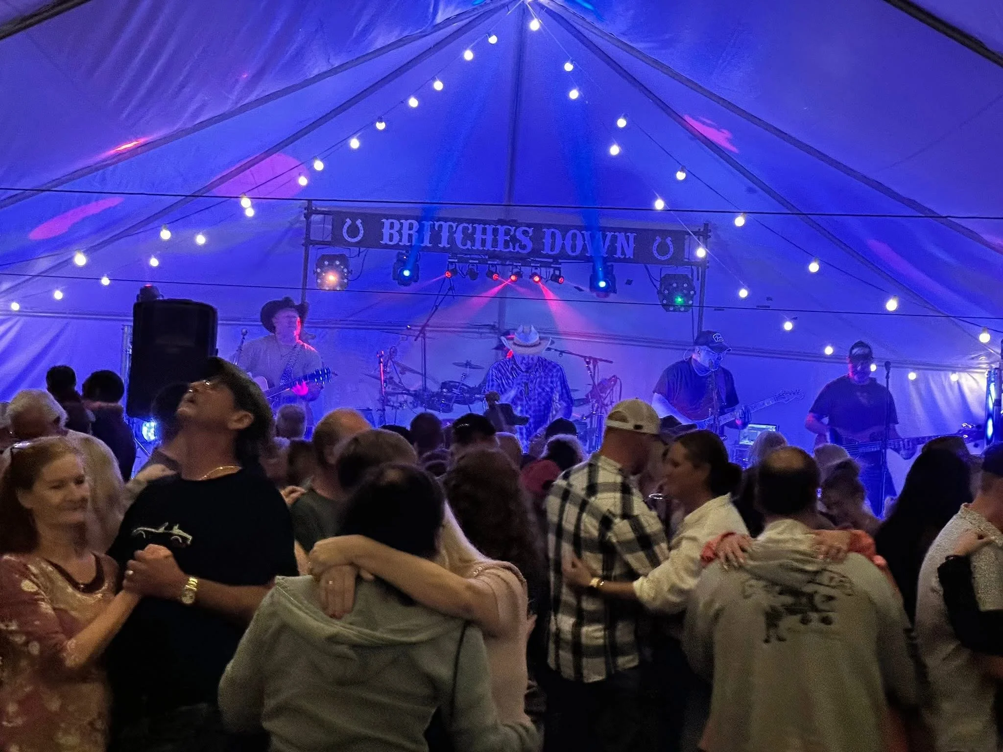 A live music concert inside a large tent with a band playing on stage under blue and purple lights, and a crowd of people dancing and enjoying the performance.