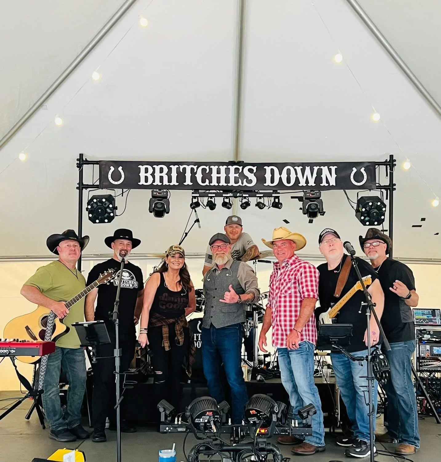 A group of musicians on stage under a tent, with a banner reading 'BRITCHES DOWN' and horseshoe decorations, performing with instruments like guitars, keyboard, and drums.