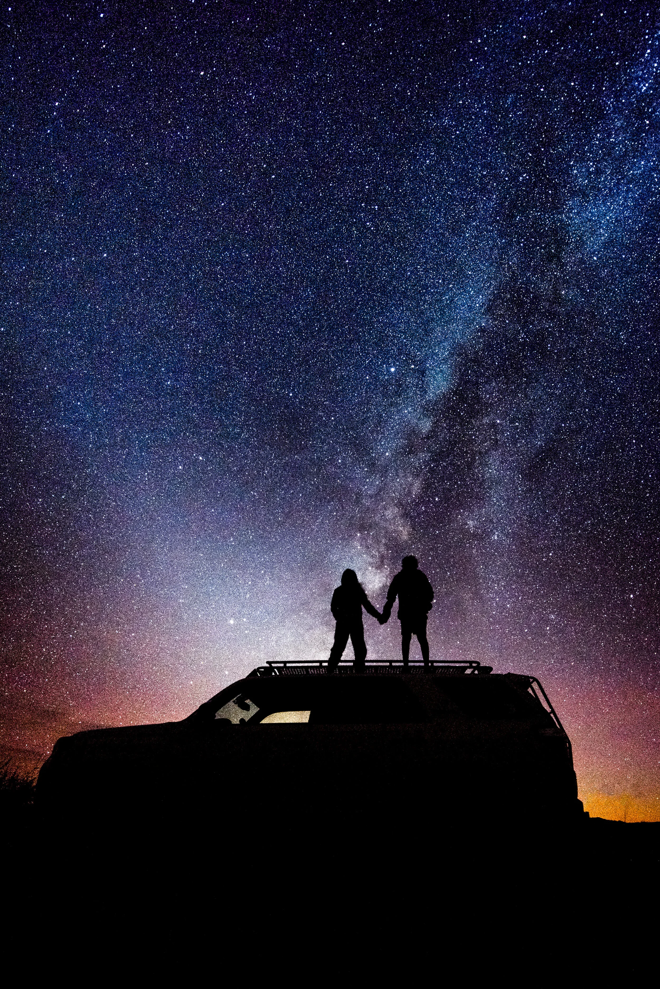 Silhouetted couple holding hands on top of a vehicle under a starry night sky with the Milky Way galaxy visible.