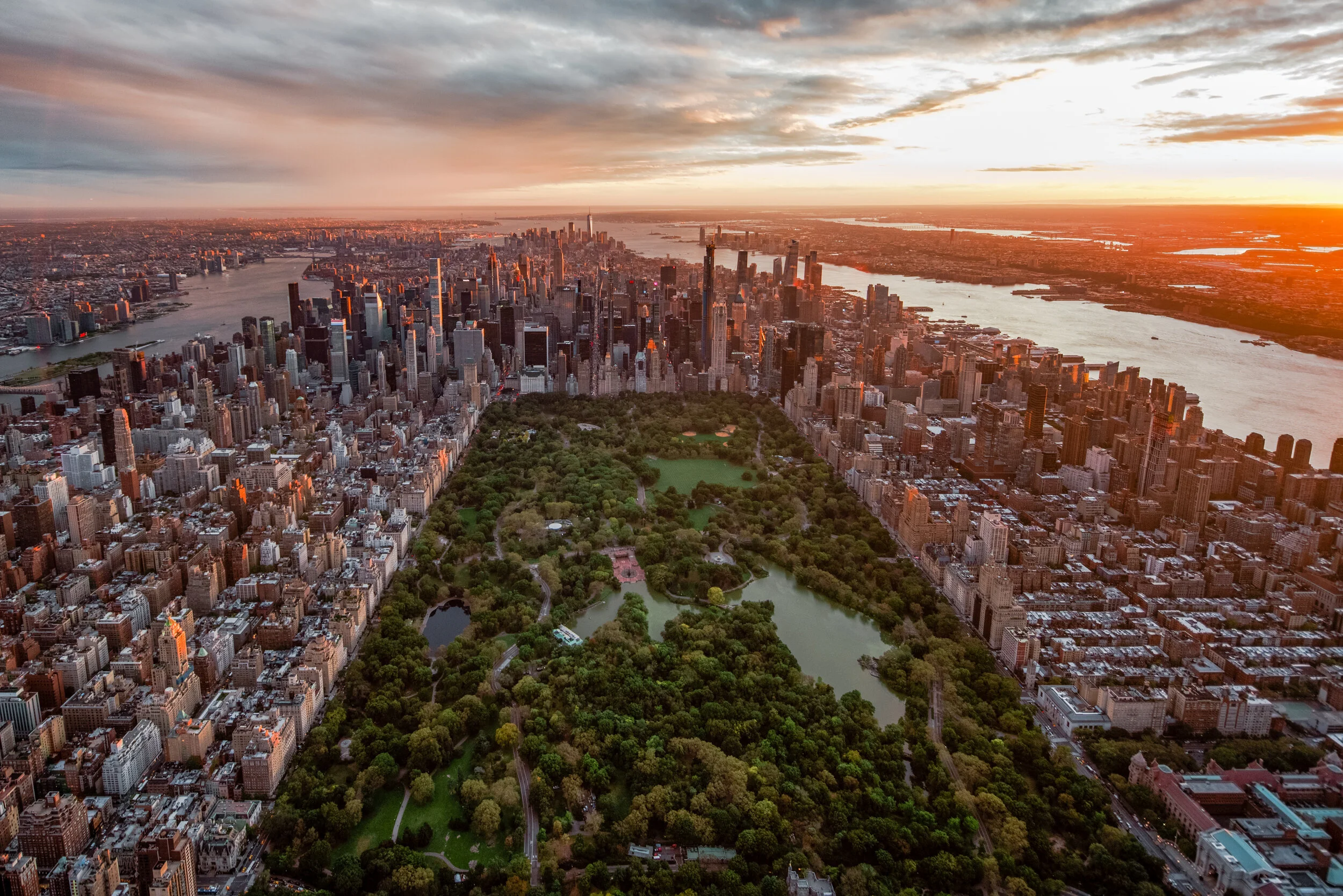 Aerial view of Central Park in New York City at sunset, with tall skyscrapers surrounding the park, the city skyline in the distance, and a river visible to the east of the park.