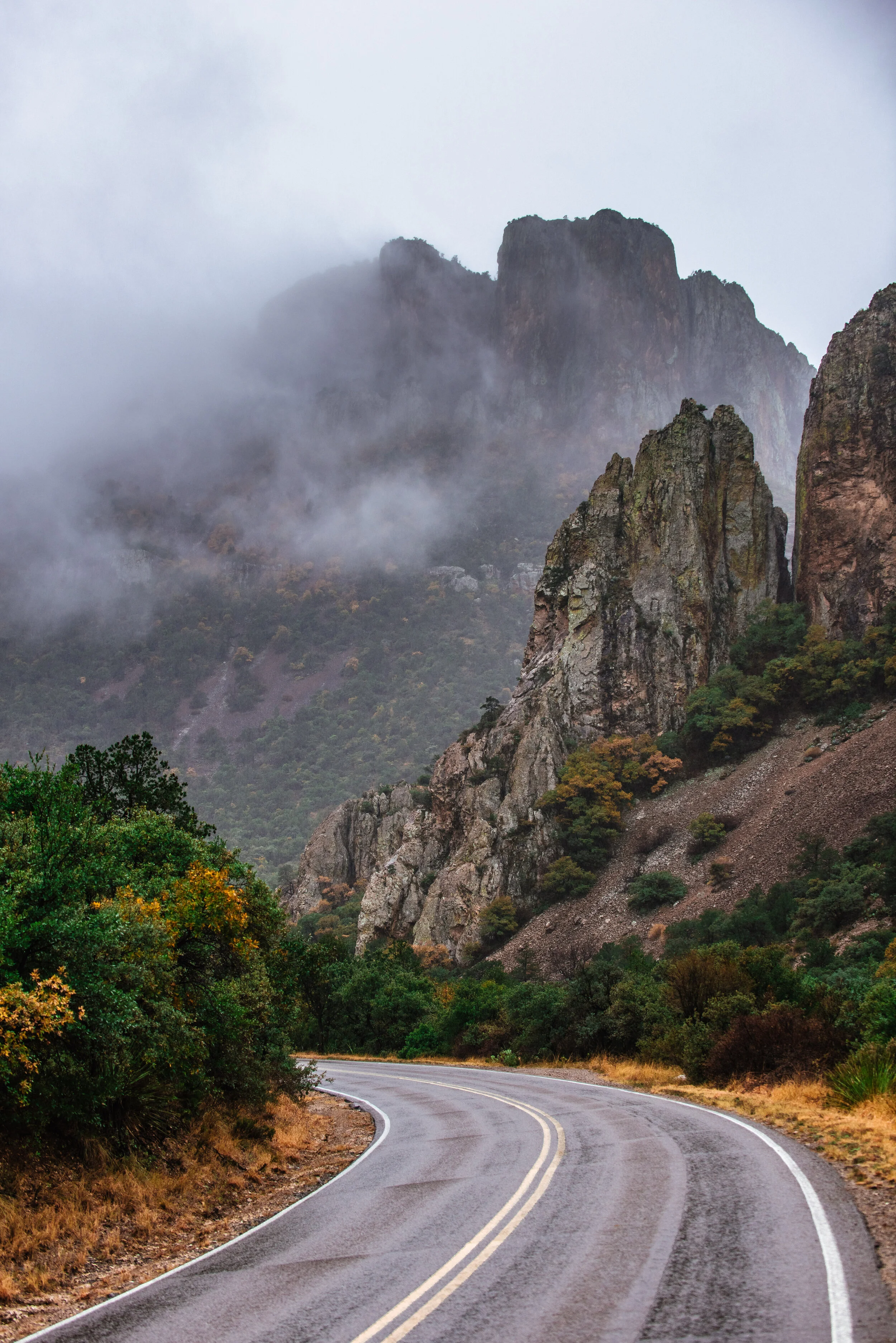A winding mountain road with green trees along the sides, rugged rocky cliffs, and fog-covered mountains in the background under a cloudy sky.