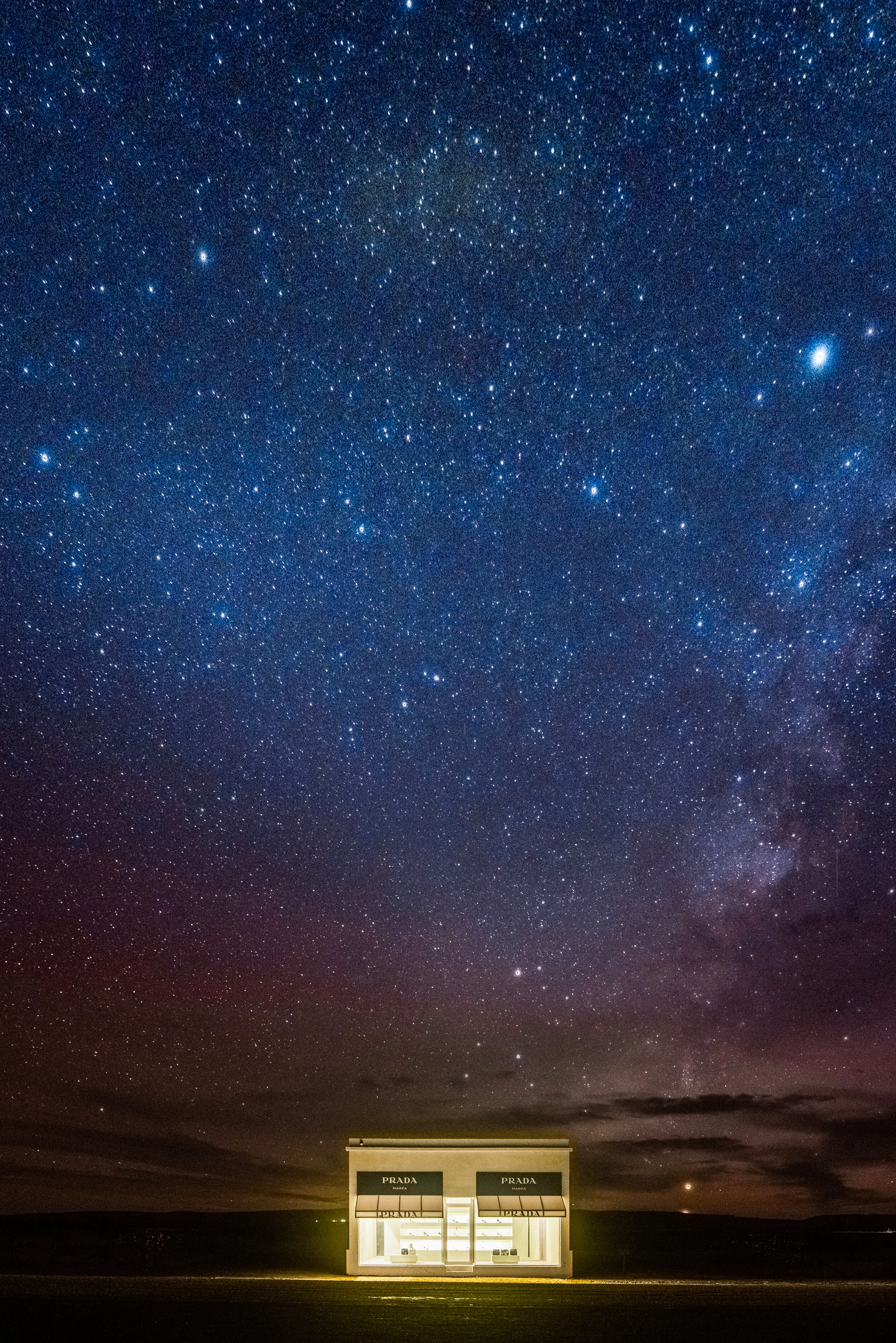 A small Prada storefront illuminated at night under a star-filled sky with the Milky Way galaxy visible.