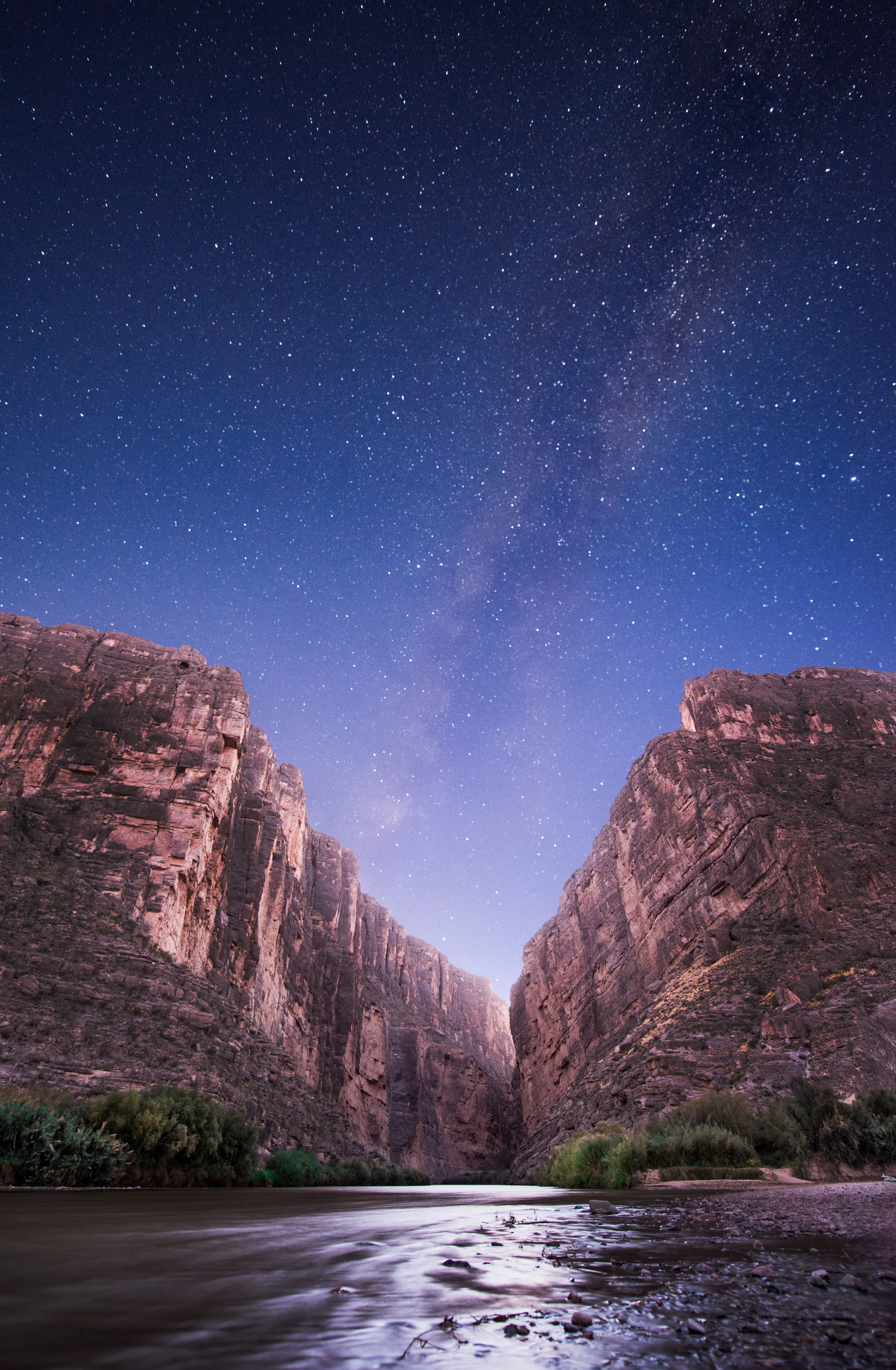 Nighttime view of a canyon with tall rock walls, a river in the foreground, and a starry sky with the Milky Way visible