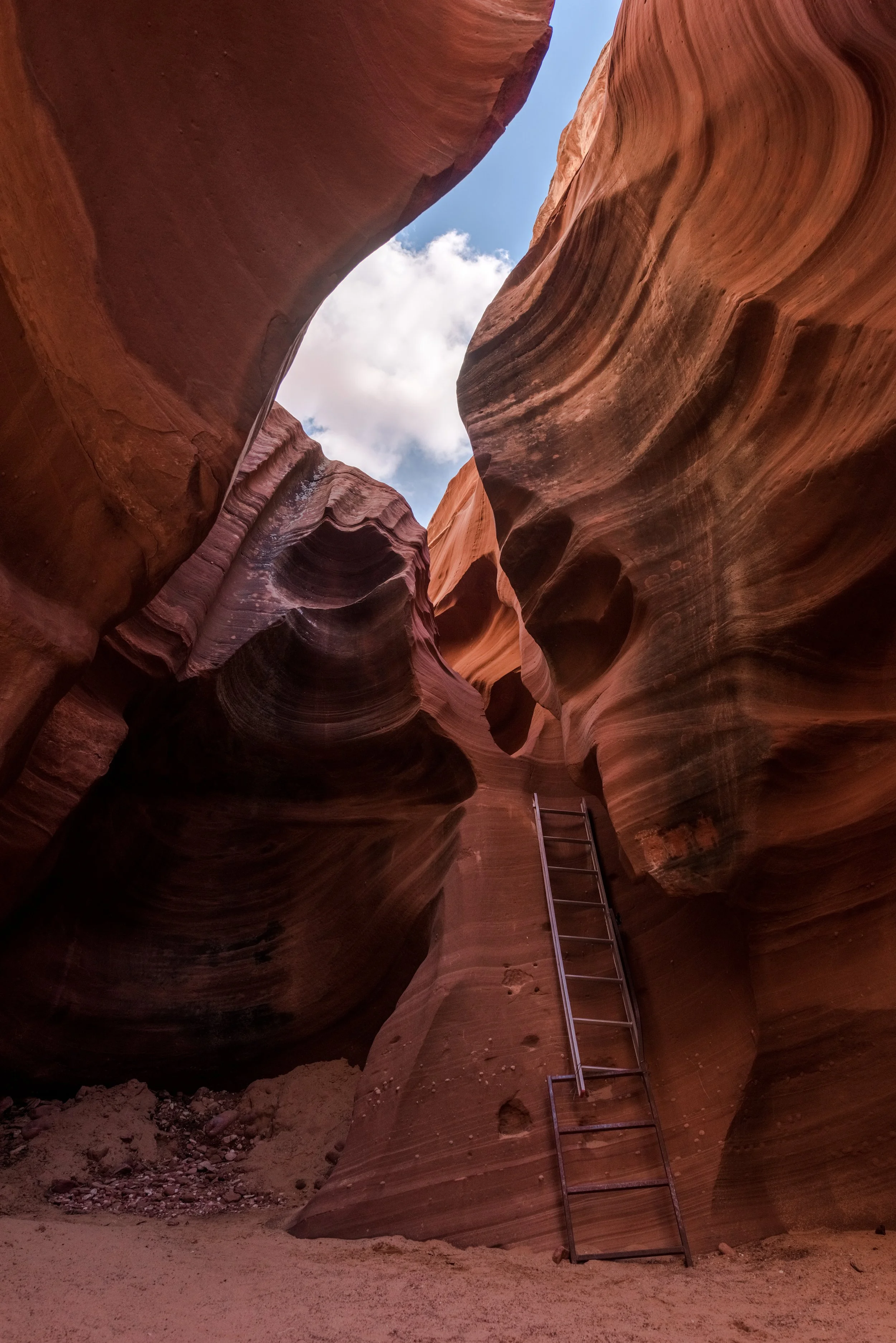 Narrow canyon with smooth, layered sandstone walls, a metal ladder leaning against one wall, and an opening to the blue sky with white clouds above.