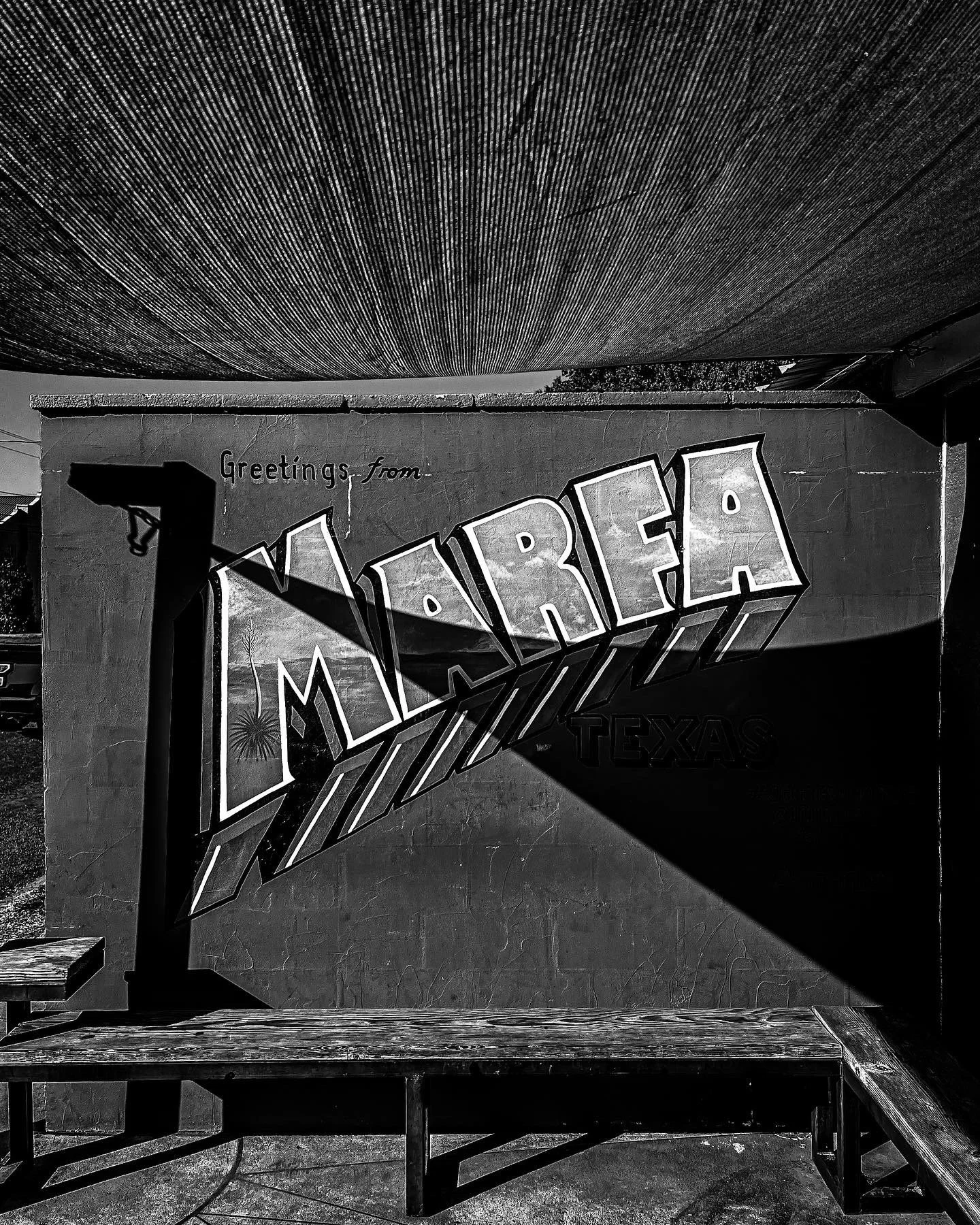 A mural on a wall reading 'Greetings from Marfa Texas' with 'Marfa' in large 3D letters and shadows, in black and white.