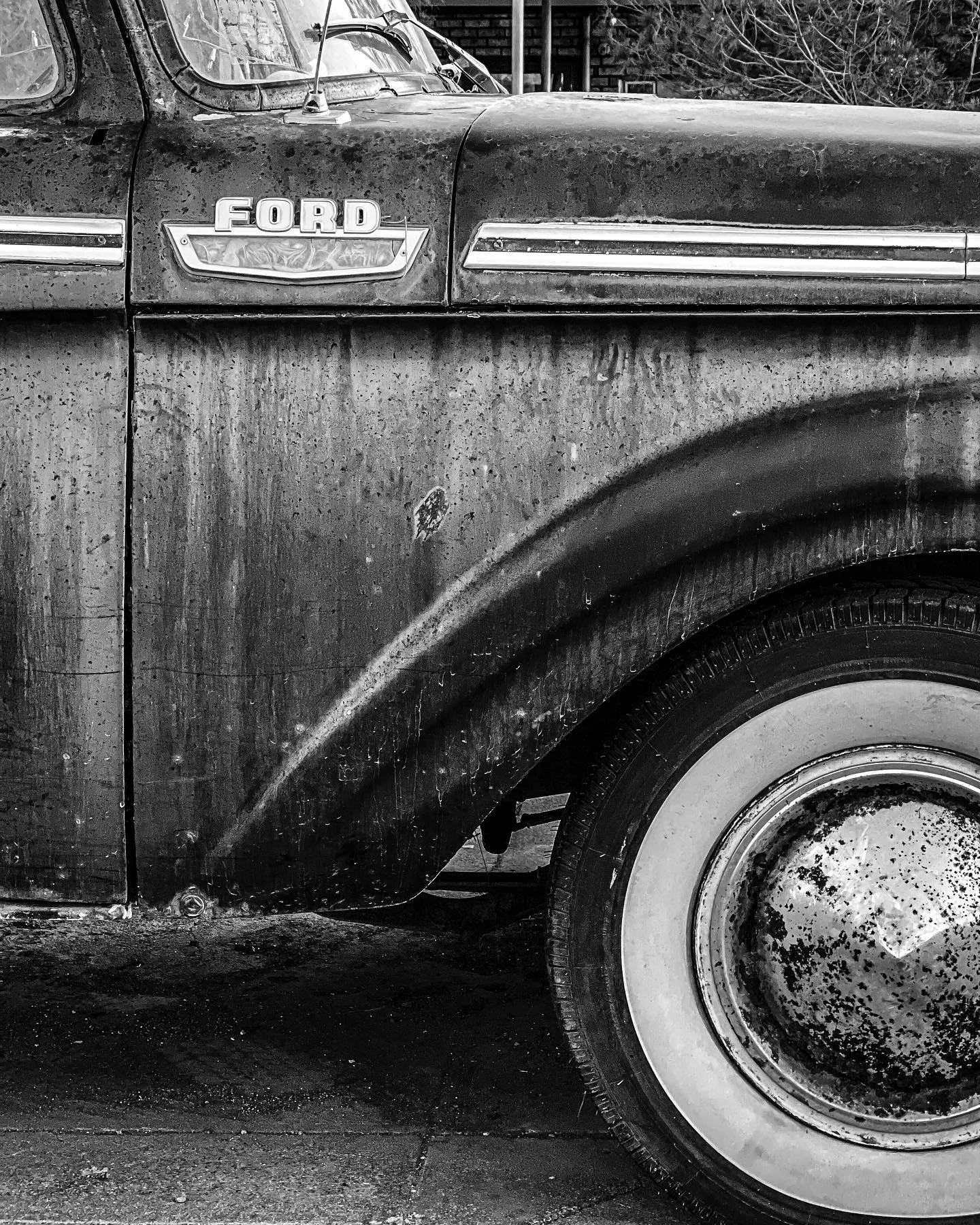 Close-up of an old, rusty Ford truck with a weathered body and a heavily corroded hubcap, black and white photograph.