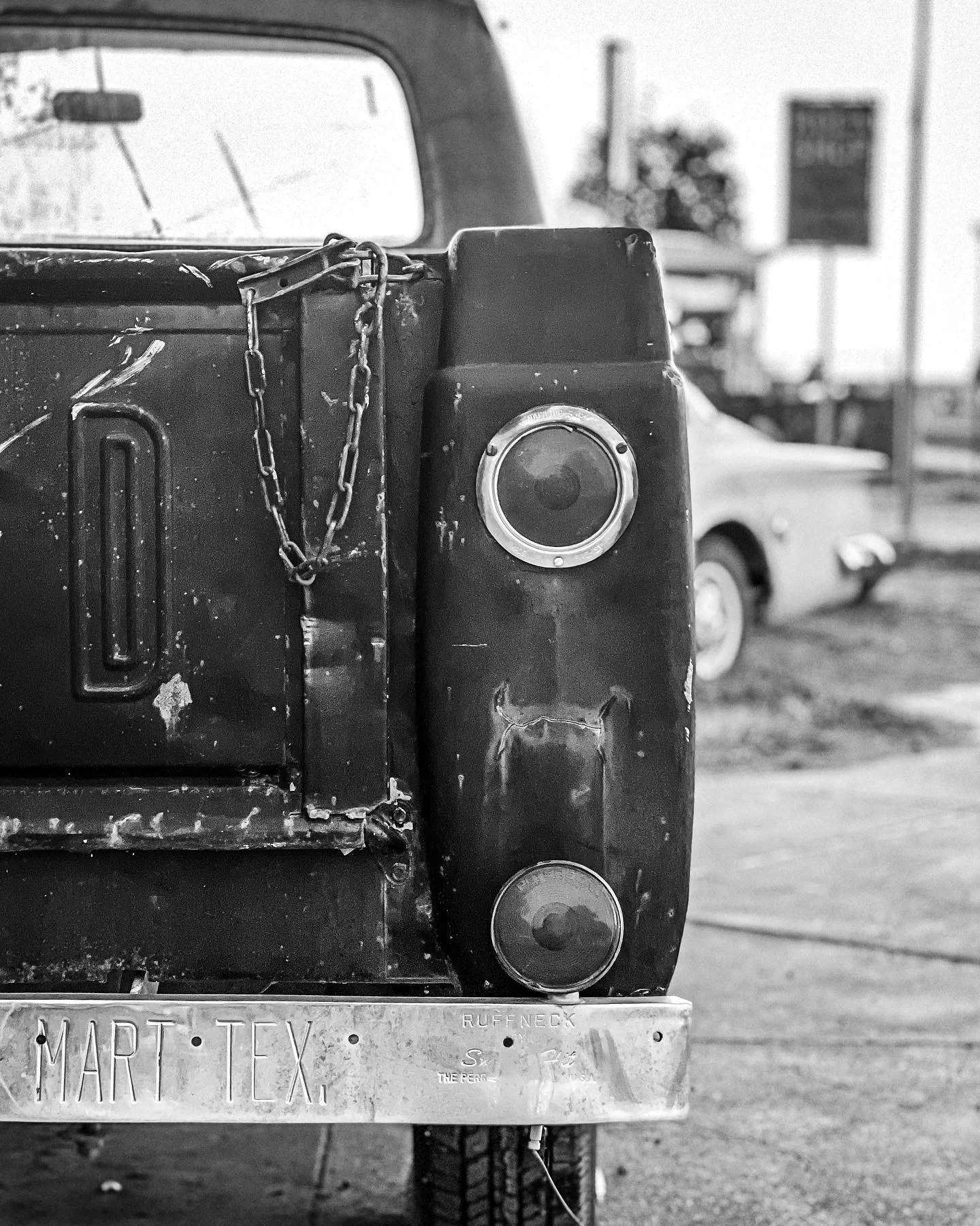 Black and white photo of the rear corner of an old, weathered pickup truck with a chain hanging from the side, parked outdoors with another vintage car in the background. The truck has a Texas license plate reading 'MART TEX'.