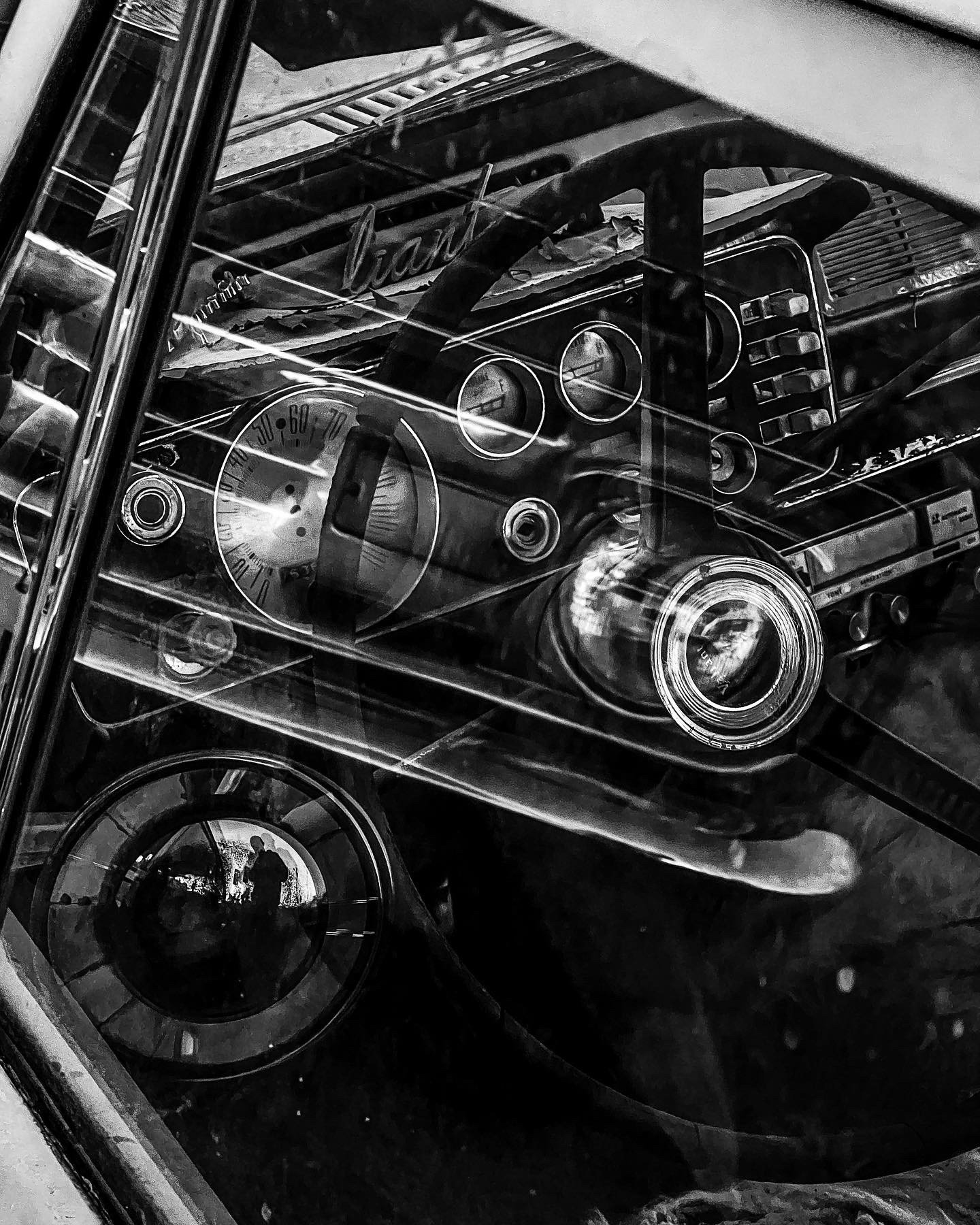 Black and white photo of a vintage car dashboard, showing a speedometer, gauges, and radio, viewed through the windshield.