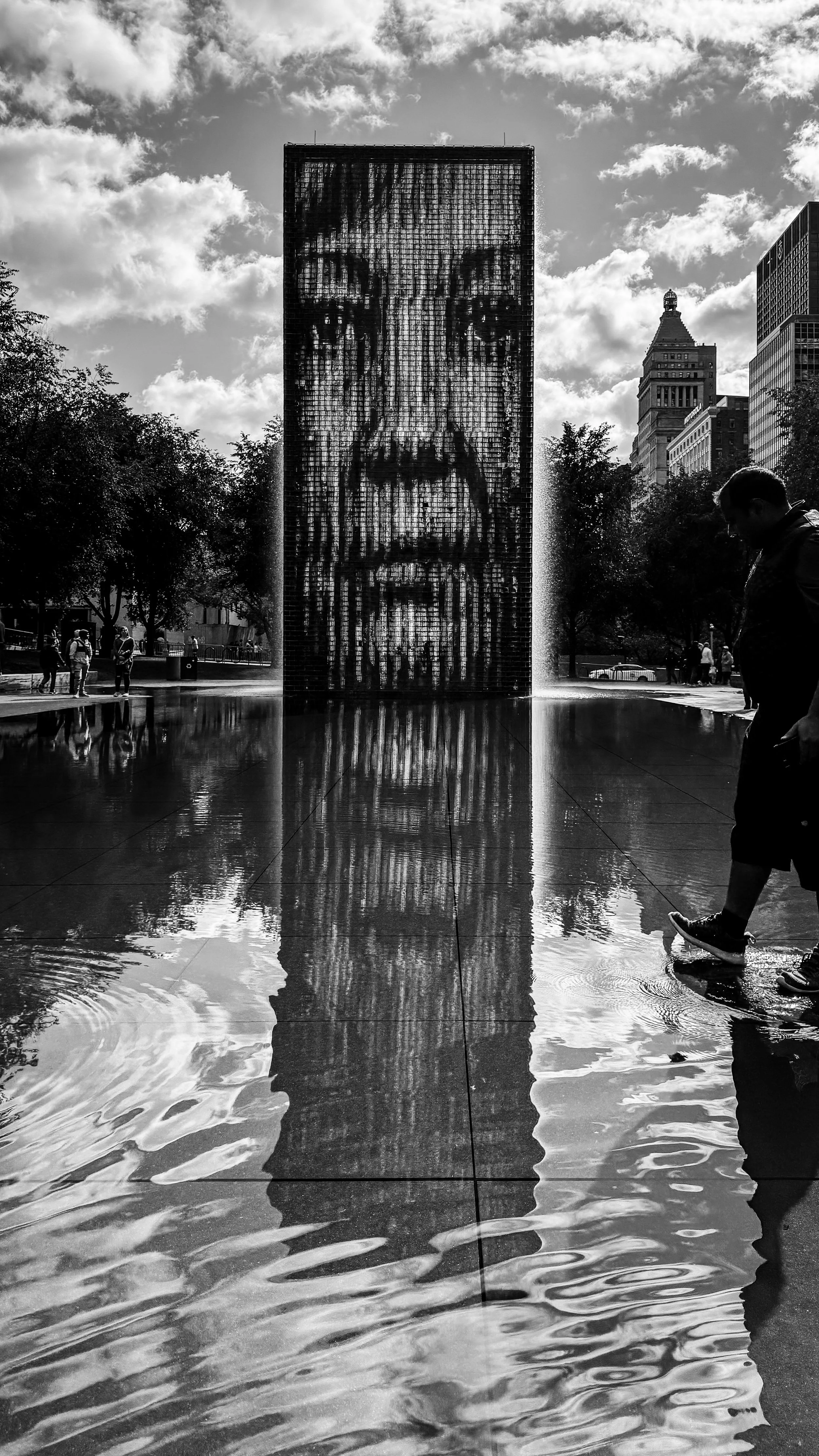 A black and white photo of a tall outdoor sculpture featuring a large pixelated portrait of a man's face. The sculpture is surrounded by water reflecting the image, with people walking nearby and some trees in the background.