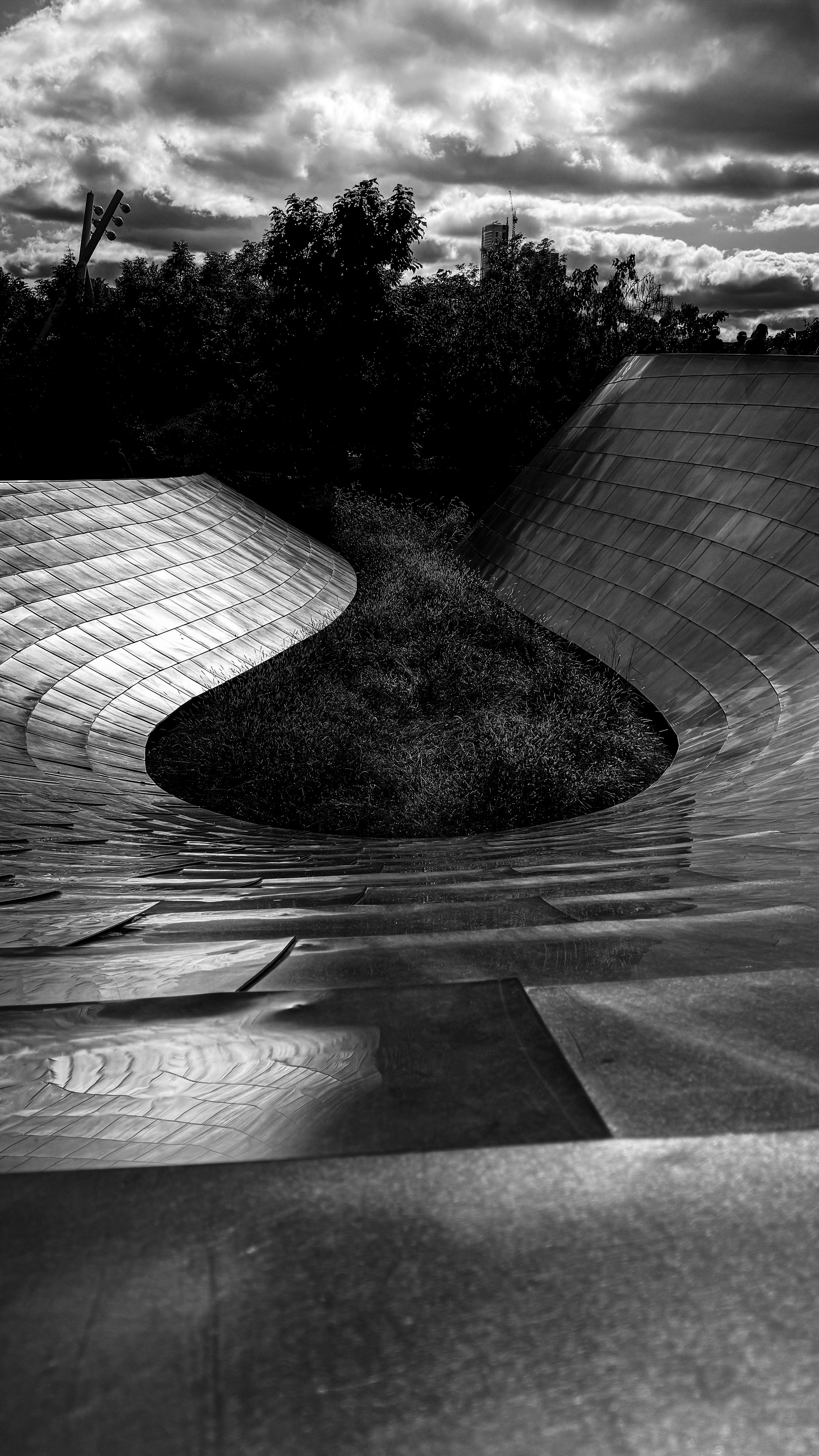 A black and white photo of an abstract, curved metallic sculpture with water splashing in the center, set against a cloudy sky and trees in the background.
