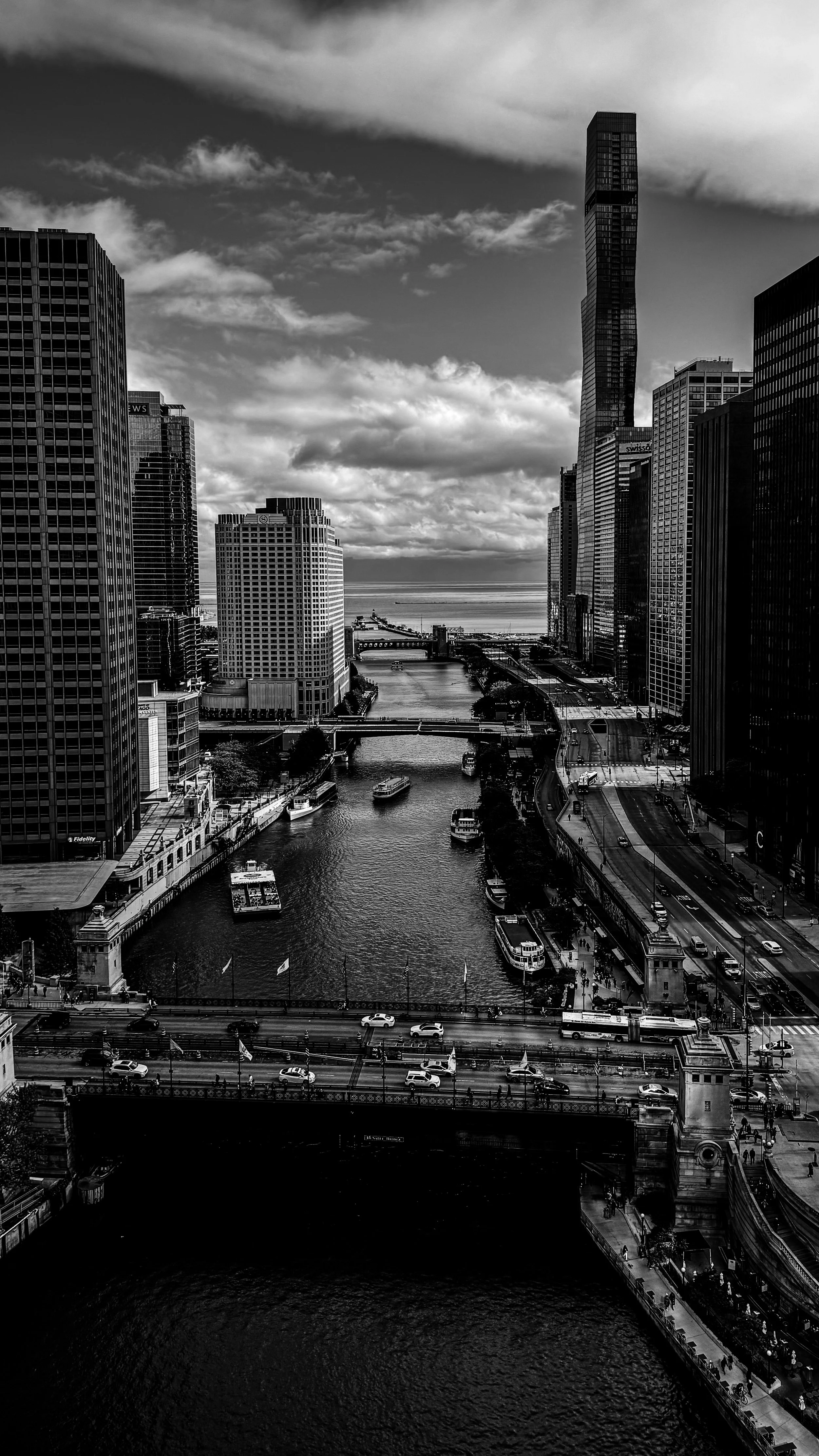 Black and white photo of a city skyline with tall buildings, a river running through the city, boats on the river, and a bridge in the foreground.