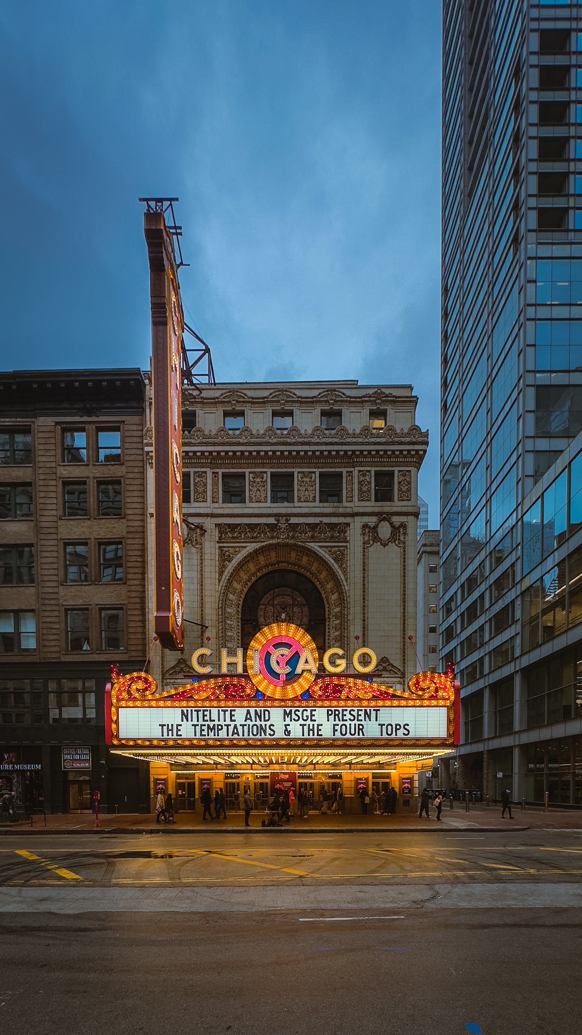 The Chicago Theatre marquee with a sign announcing a concert featuring Nitelite, MSGE, The Temptations, and The Four Tops, located on a city street at dusk.