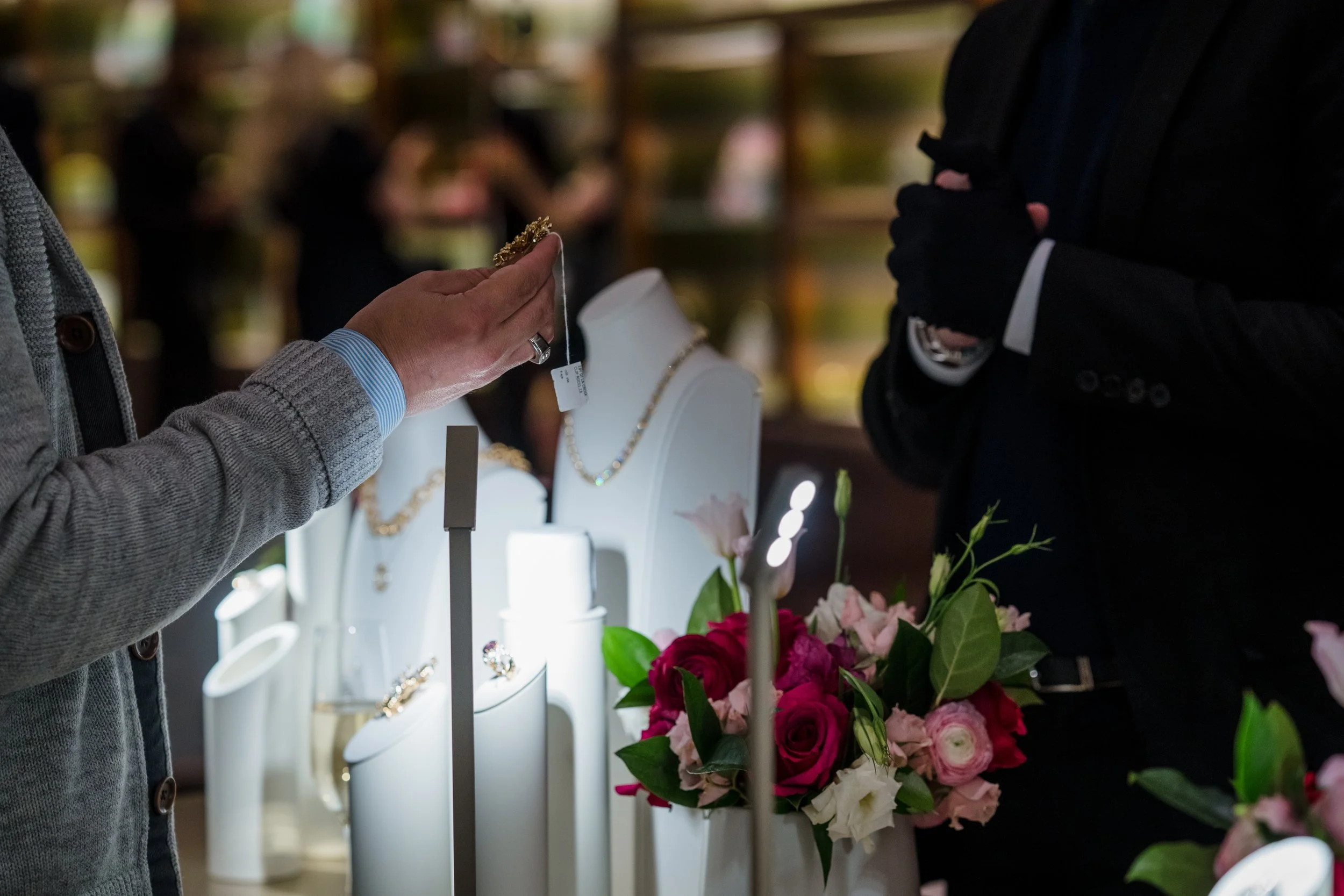 Two men are examining jewelry, with a display of necklaces and rings, and a bouquet of pink and red flowers nearby.