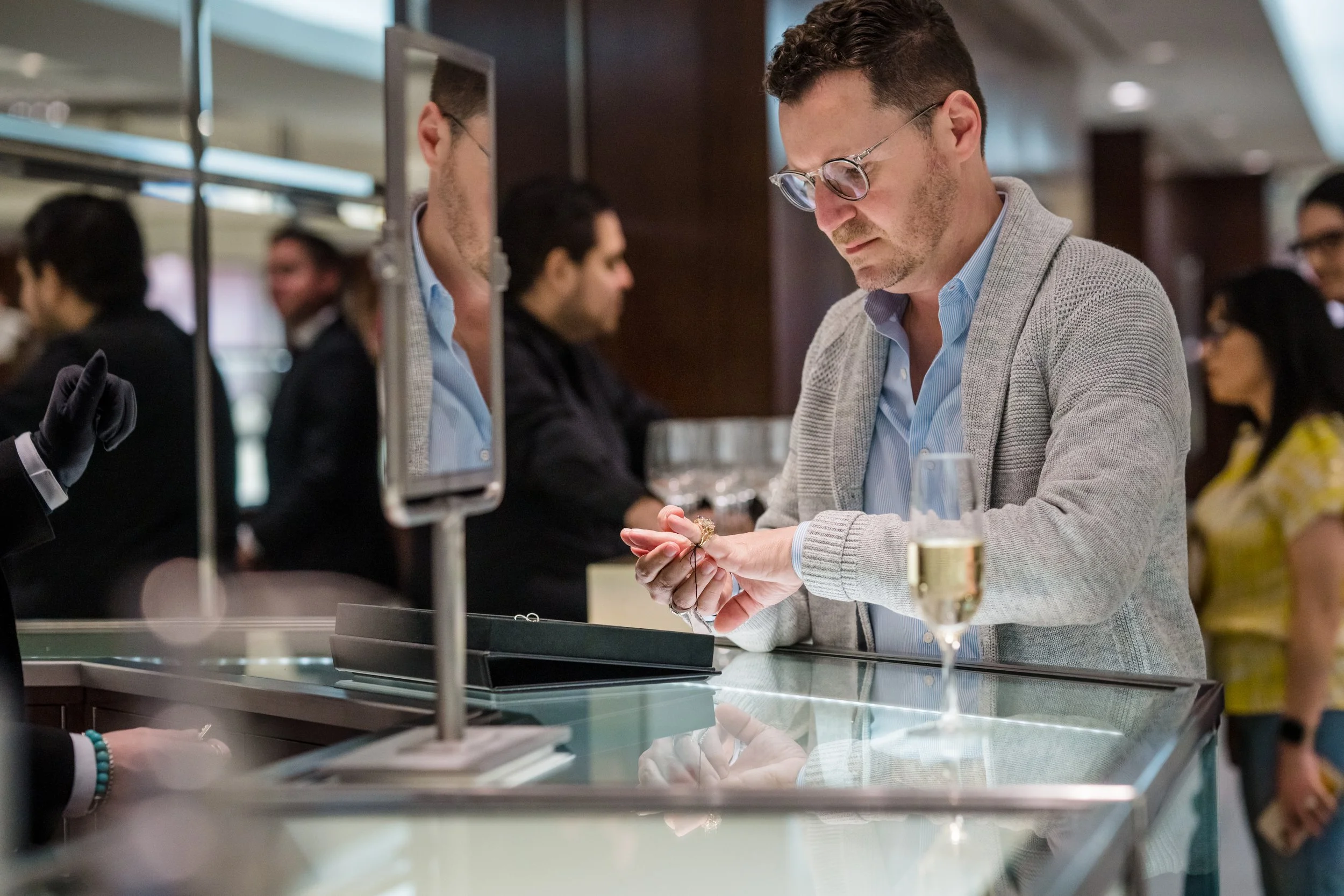 A man with glasses and a gray cardigan examines jewelry at a store counter, with a champagne glass on the counter, in a busy shopping mall or jewelry store.